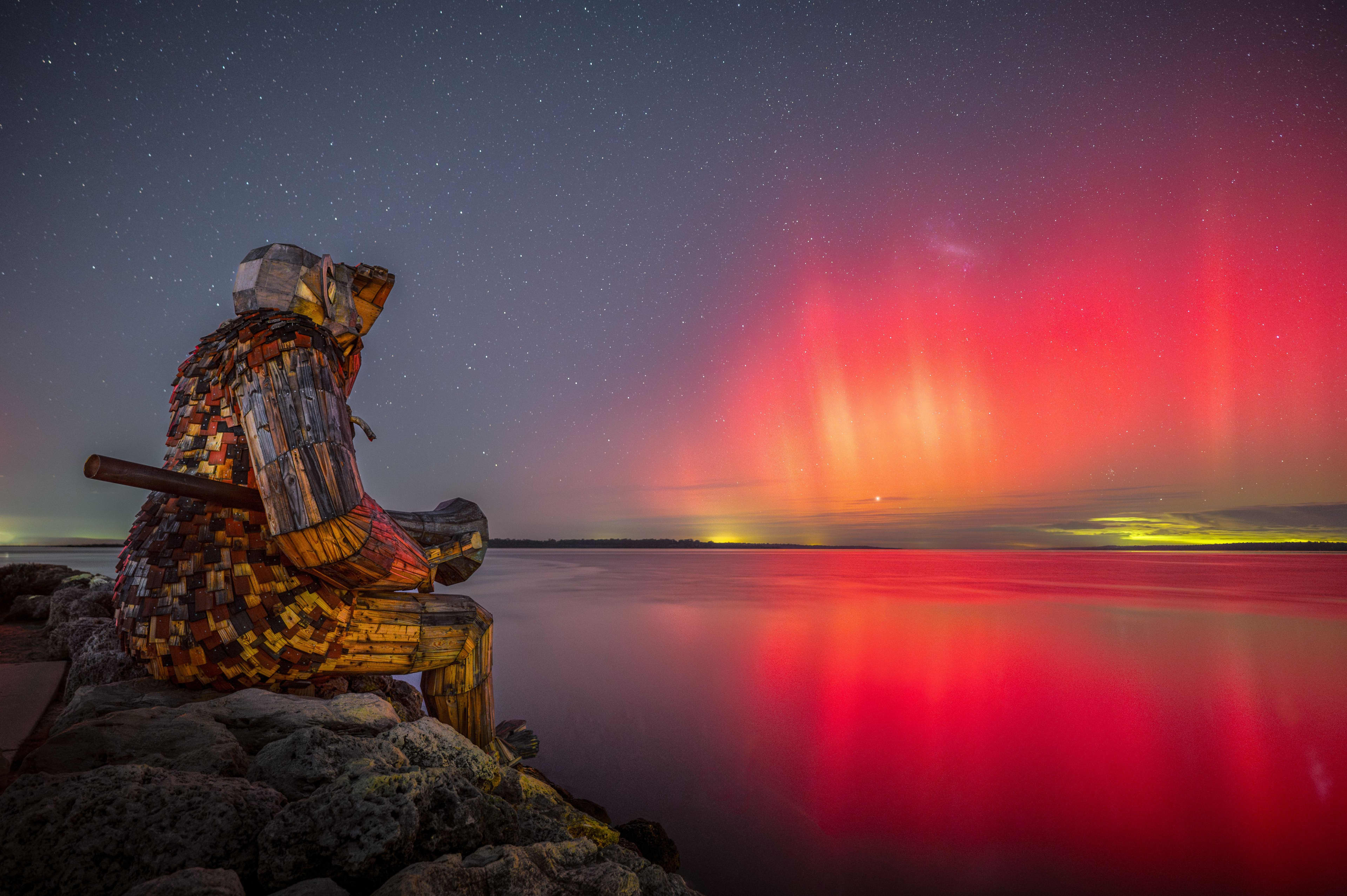 An astro image of a large wooden troll sculpture overlooking an estuary with the sky lit up with an aurora.