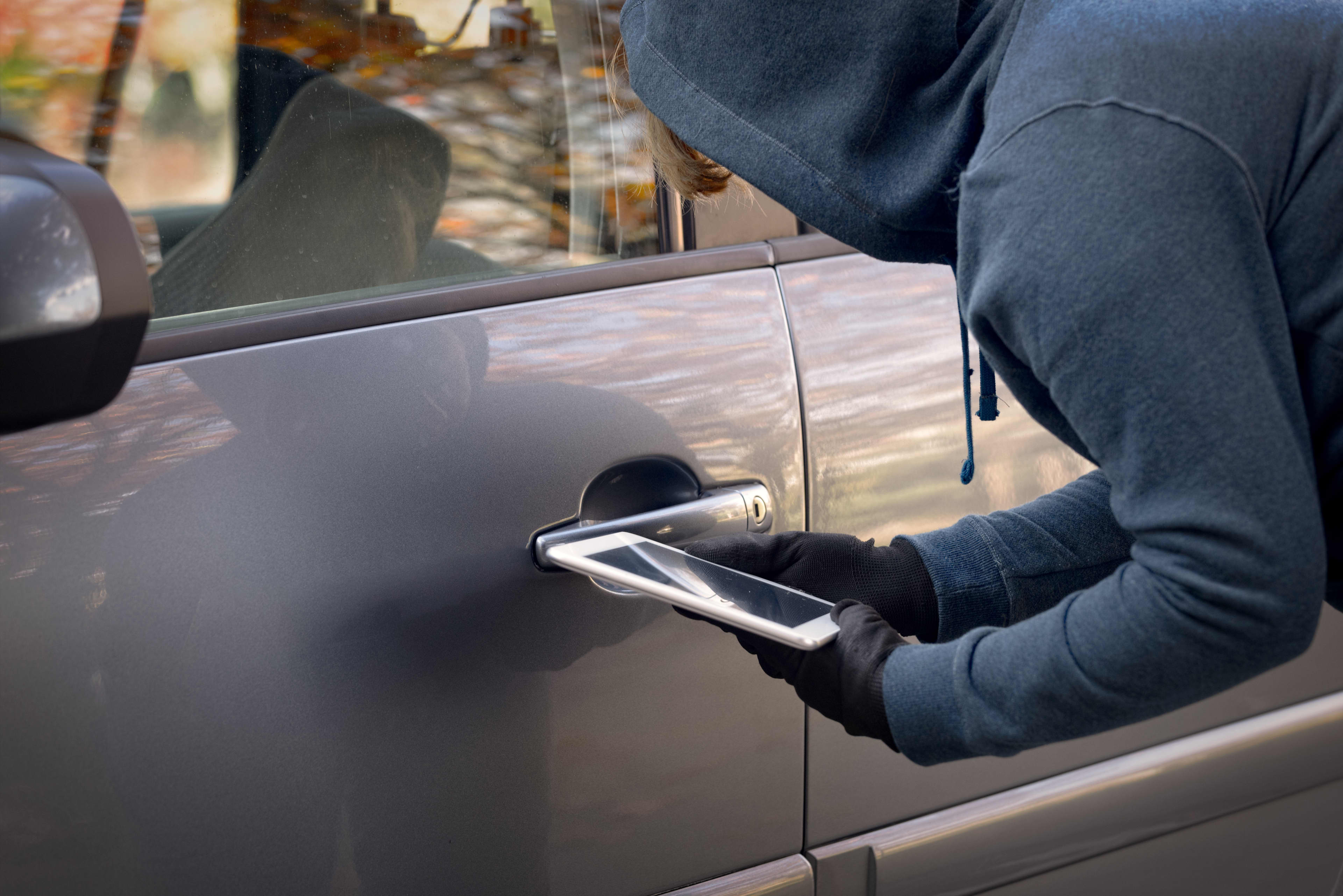 A hooded thief holds a tablet device near a car door