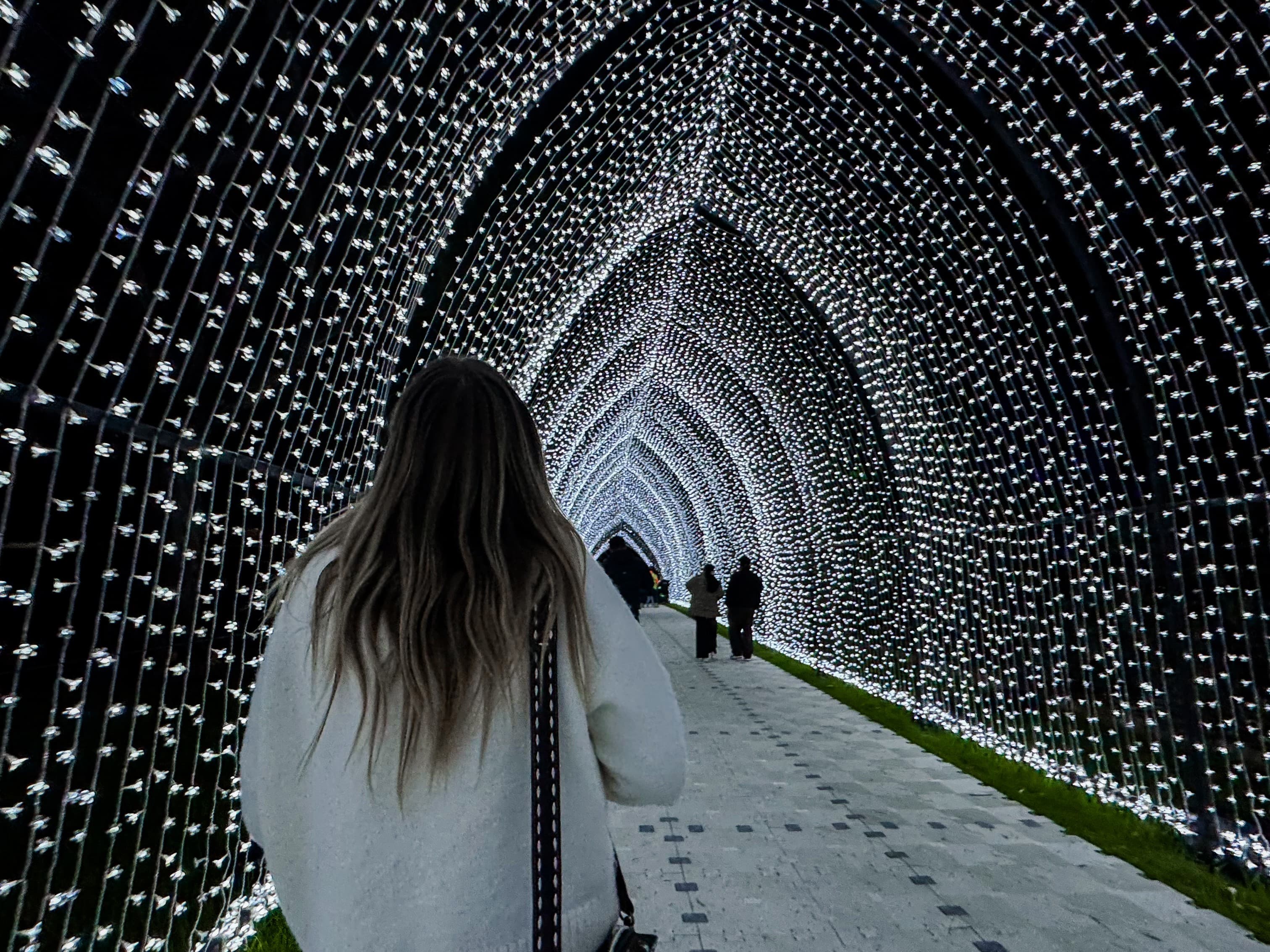 Person walking through an arched tunnel of white lights at night, with illuminated strands forming a glowing canopy overhead and other visitors visible in the distance.