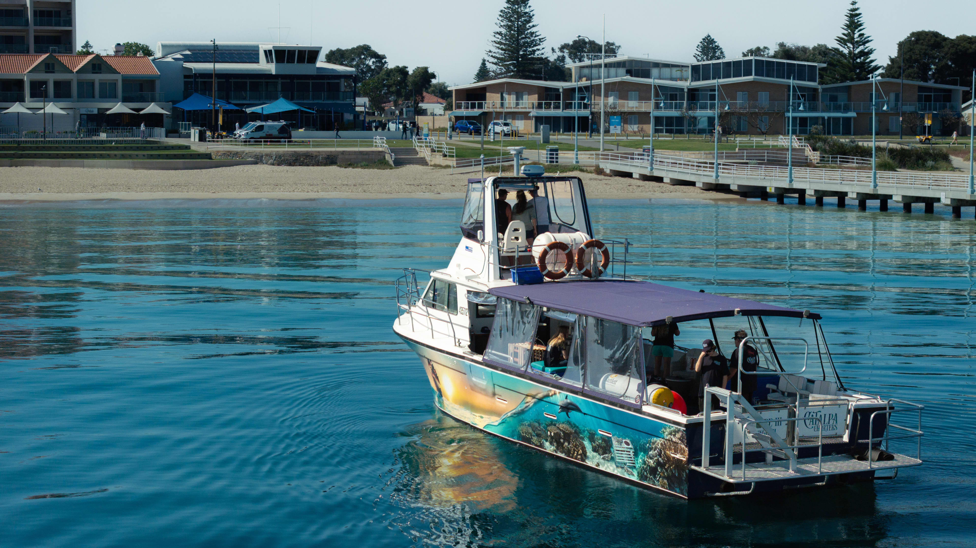 A small tour boat with a colorful ocean-themed mural on its side moves through calm blue water near a waterfront. Several people are on board under a canopy, and life rings are mounted on the upper deck. In the background, there are modern low-rise buildings, a pier with railings, and a sandy shoreline lined with trees.
