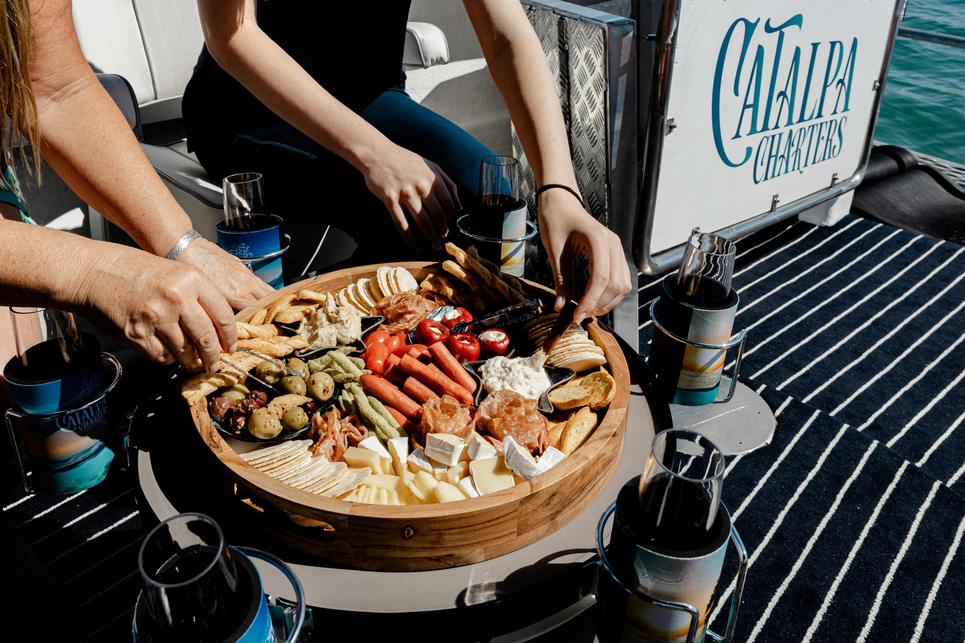 A close-up shot shows a variety of hands reaching for food from a large, round wooden grazing platter. The platter is filled with an assortment of snacks, including crackers, sliced meats, olives, various cheeses, and dips. The scene is set on a boat, indicated by the "Catalpa Charters" logo on a white panel in the background and the blue and white striped floor. Several glasses of dark liquid in insulated holders are placed around the table.