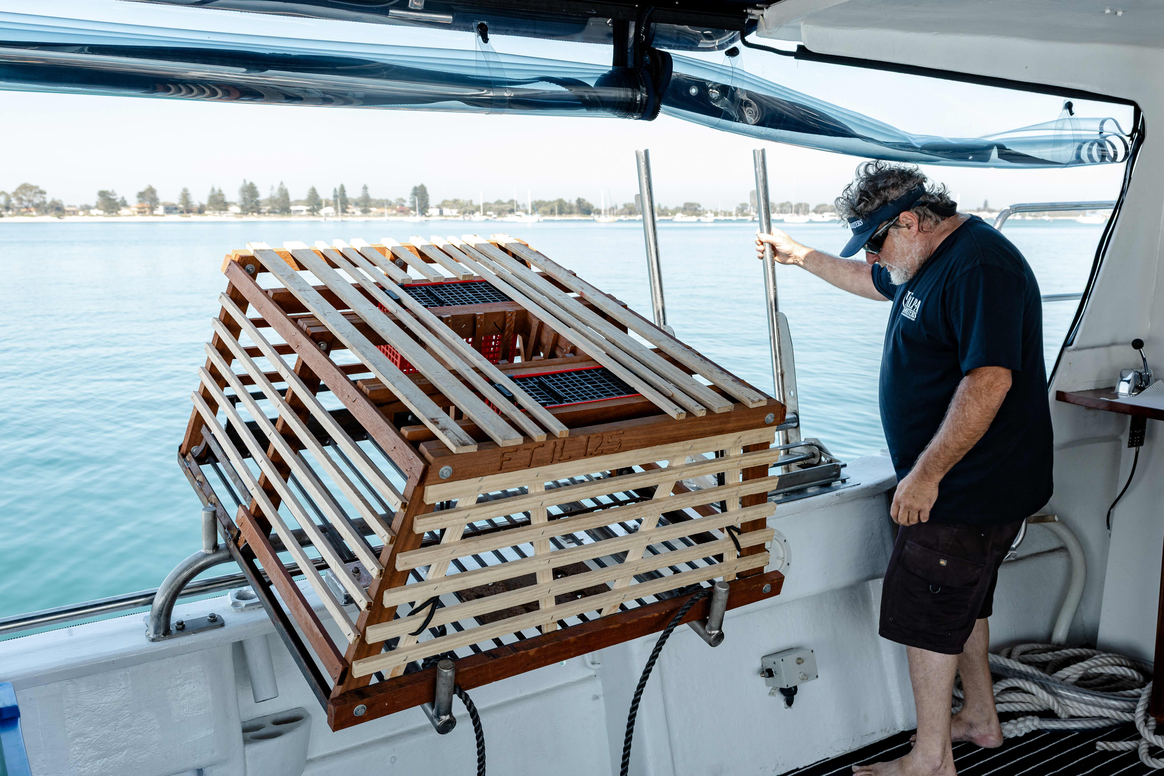 A man stands on a boat, positioning a large wooden lobster trap over the side into the blue water. The trap is a slatted, rectangular structure with a rope attached to its base. The man is wearing a dark t-shirt, shorts, and a visor, and a distant shoreline with buildings and trees is visible across the water.