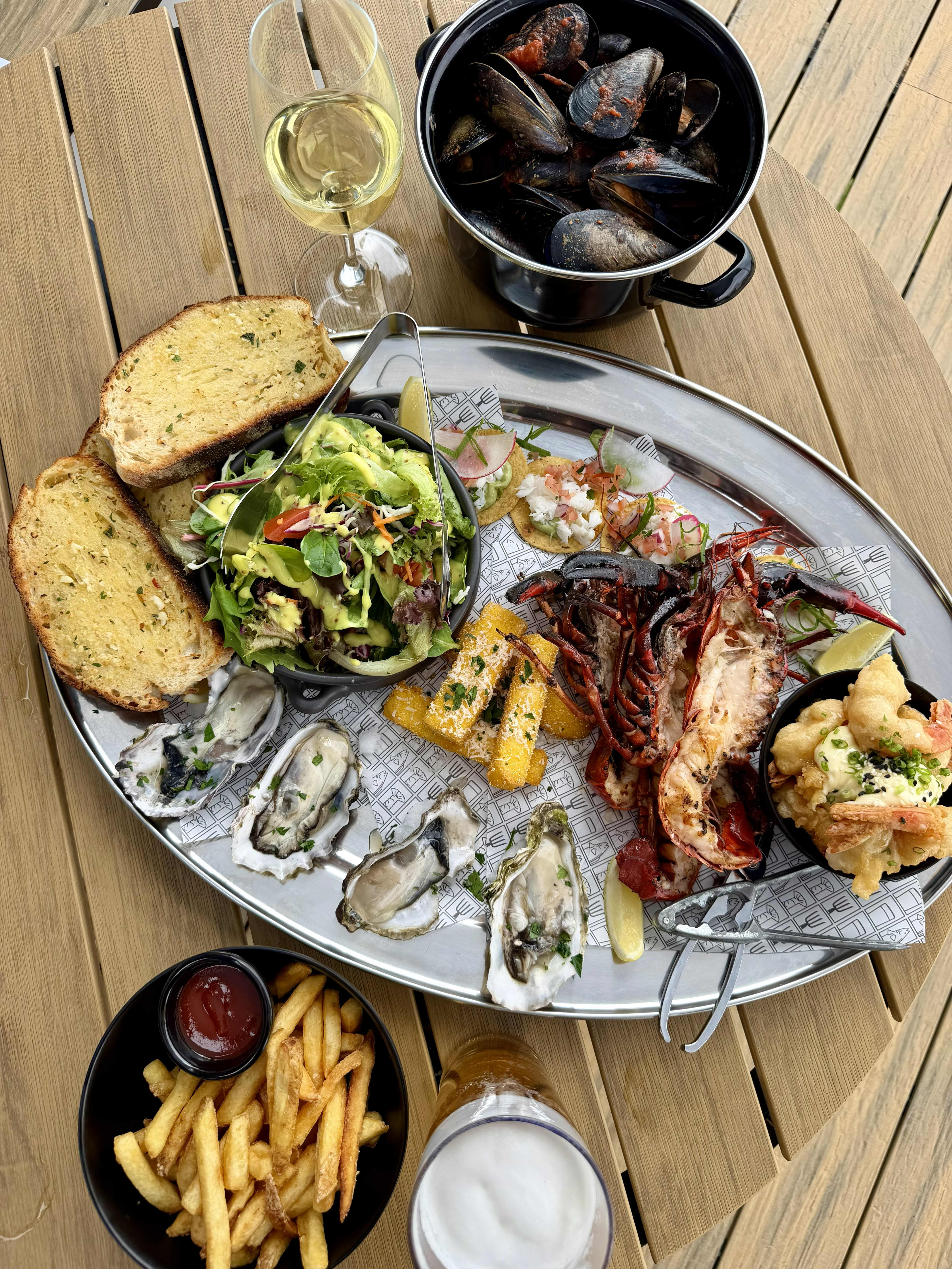A top-down view of a seafood platter served on a large oval silver tray, set on a wooden table. The platter includes split grilled marron (crayfish), four fresh oysters on the half shell, a bowl of green salad, crispy polenta chips, slices of toasted garlic bread, and small seafood bites on crackers. Surrounding the tray is a black pot filled with steamed mussels, a bowl of french fries with ketchup, a glass of white wine, and a glass of beer.