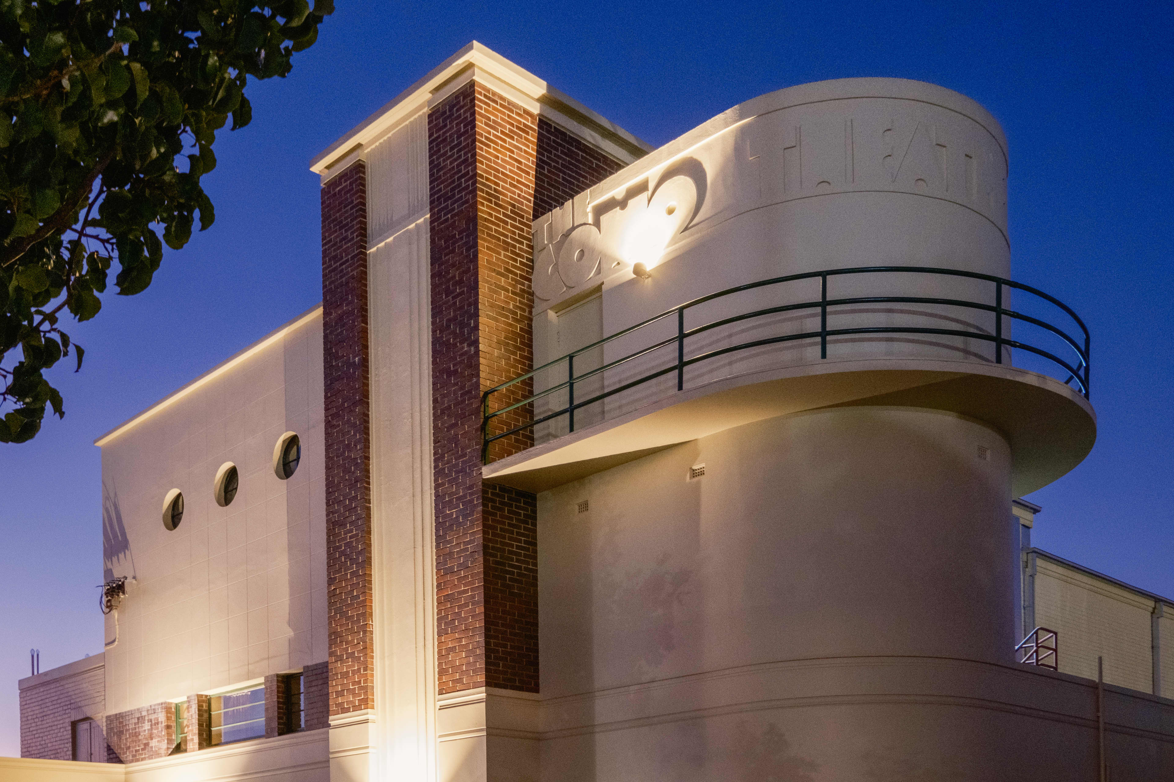 An Art Deco-style building illuminated at night against a deep blue sky. The structure features a prominent, curved white concrete balcony resembling a ship's bow, accented by dark metal railings. A tall, vertical pillar made of red brick and cream trim anchors the center of the facade, while a row of three small, round porthole windows sits on the upper left wall.