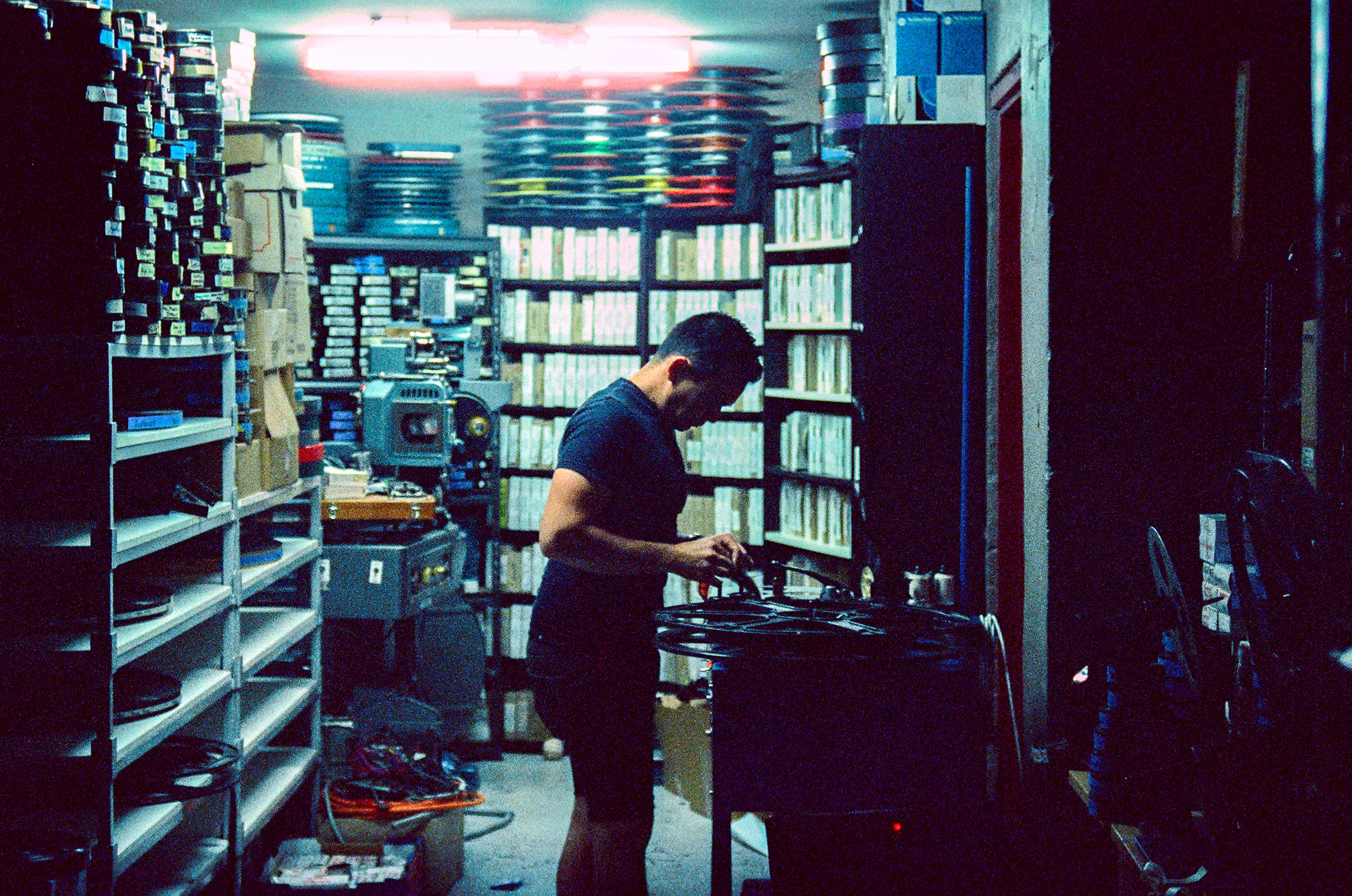 A man stands in profile inside a dimly lit, cluttered film archive or projection room, working with a large film reel on a table. The room is densely packed from floor to ceiling with shelves holding countless film canisters, boxes, and media cases. A bright red neon light fixture on the ceiling illuminates the upper background, contrasting with the cool, dark tones of the foreground.