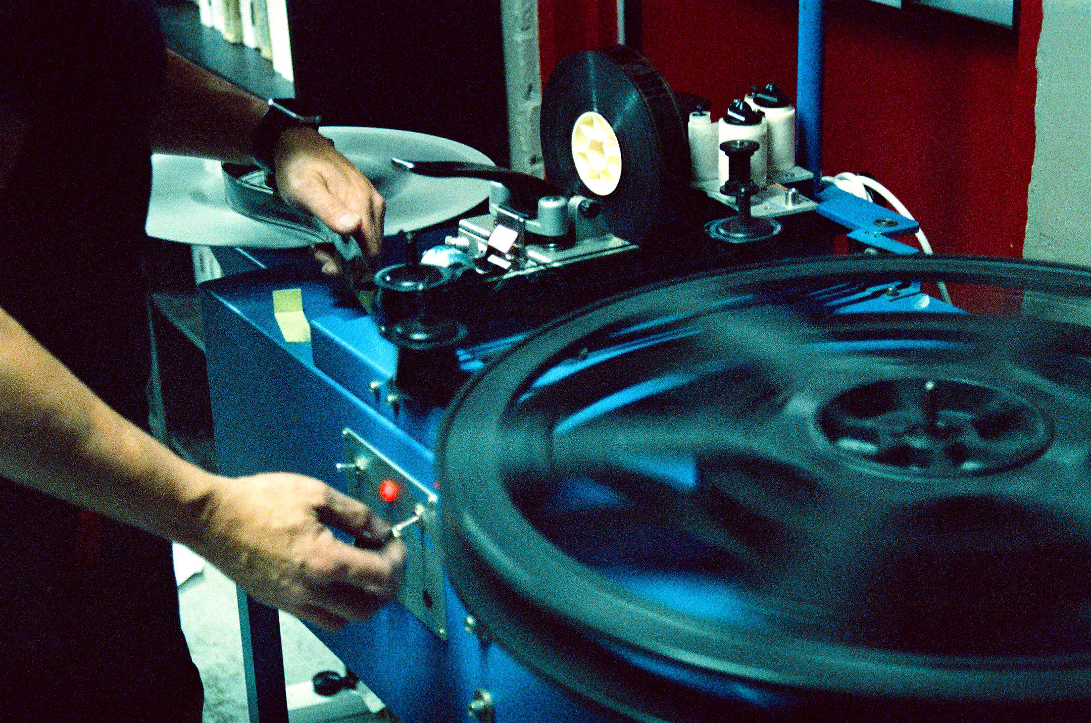 A close-up shot focuses on a person's hands operating a blue film inspection or editing machine. In the foreground, a large film reel spins rapidly, creating a motion blur. The operator's left hand adjusts a toggle switch next to a glowing red indicator light on the machine's control panel, while their right hand guides the film strip near a smaller, vertical reel mounted on top.
