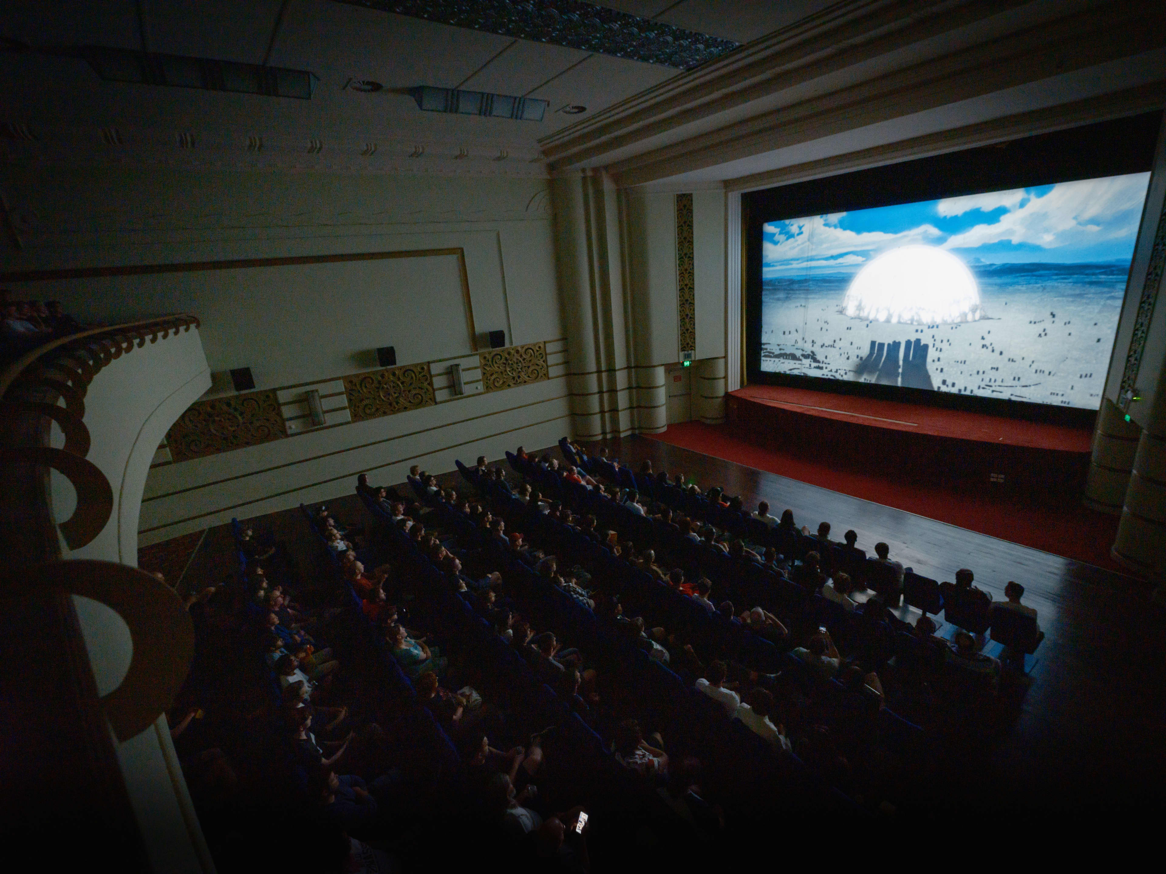 A high-angle view from a balcony inside a historic movie theater filled with an audience watching a film. A large, bright screen at the front displays an animated scene featuring a large white dome or sun on a horizon under a blue, cloudy sky. The theater's interior features classic architectural detailing, including a grand proscenium arch framing the screen and decorative panels along the side walls.