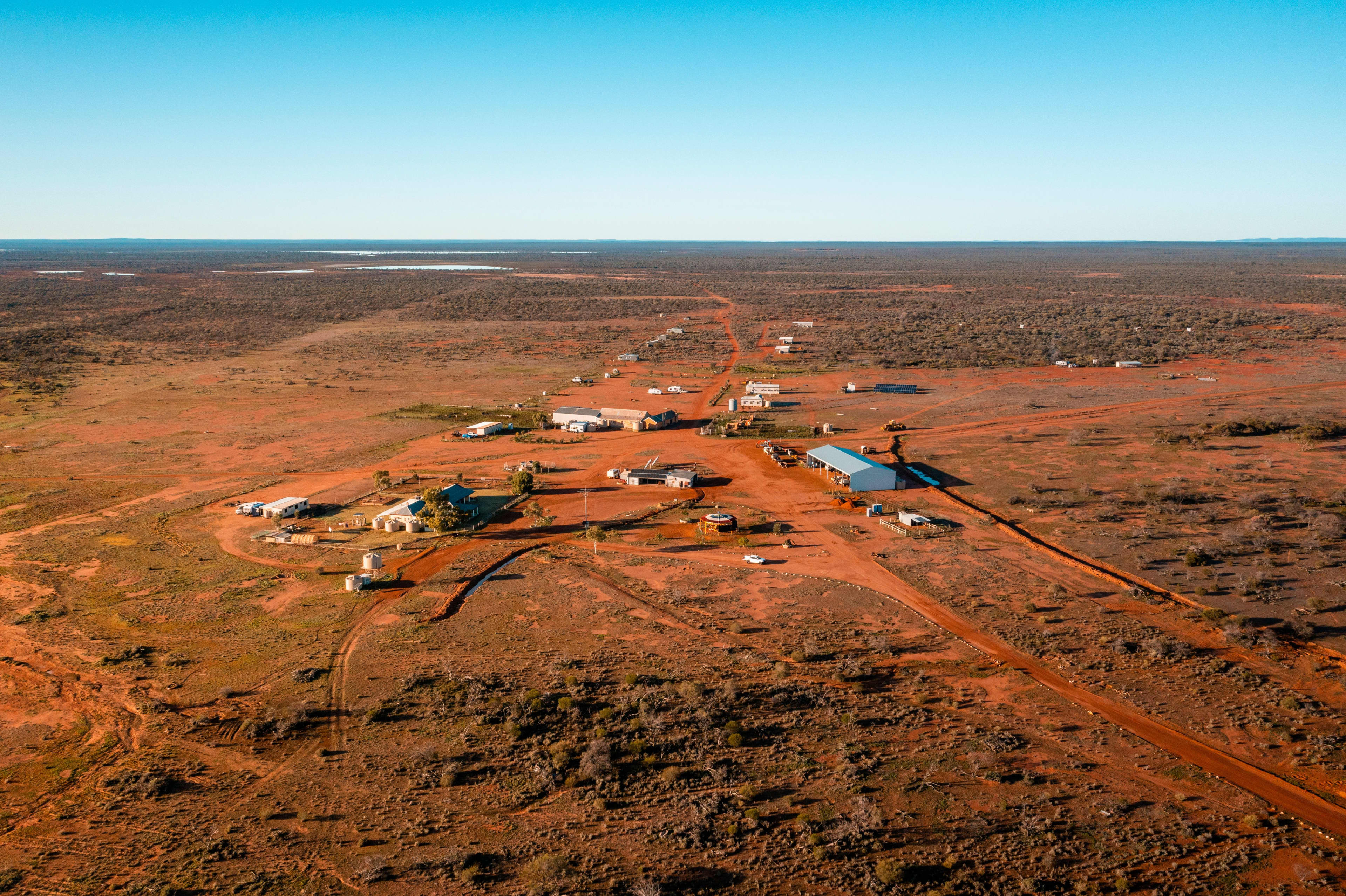 Aerial view of a remote desert settlement with scattered buildings connected by red dirt roads, surrounded by vast flat scrubland under a clear blue sky. Small water bodies are visible in the distance, emphasizing the isolated landscape.