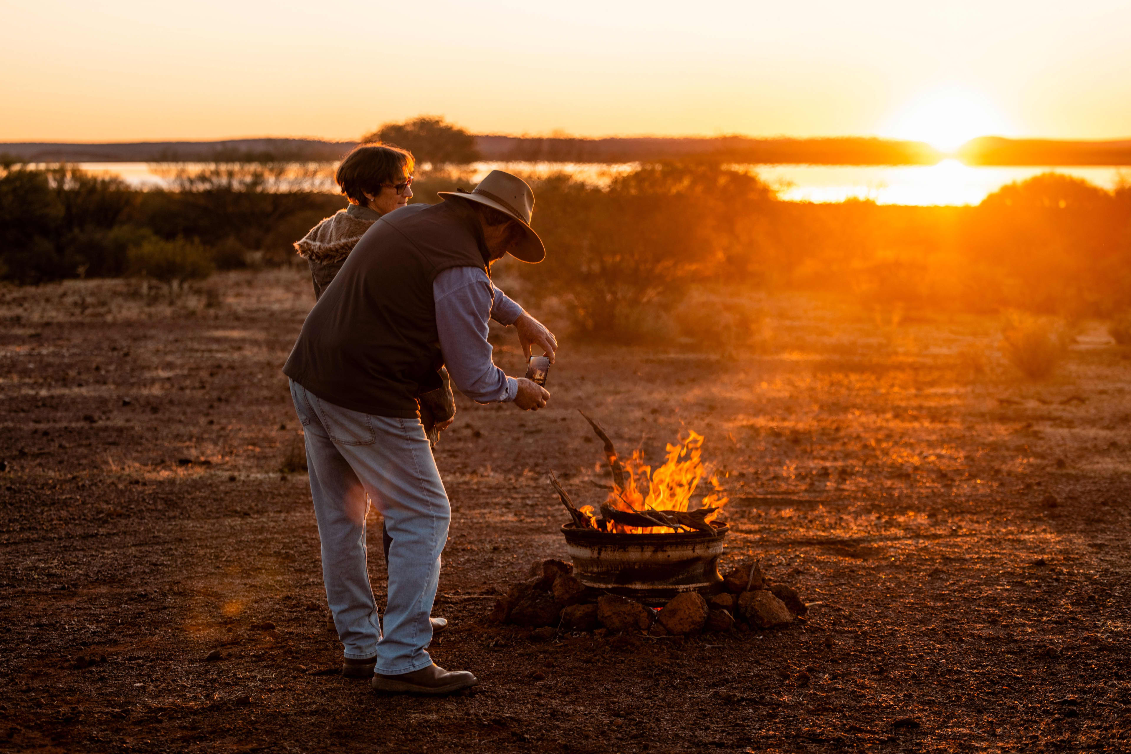 Two people stand beside a small campfire at sunset in a dry, open landscape. One person wearing a wide-brimmed hat pours a drink while the fire glows in the foreground, with the sun setting over a lake and silhouetted shrubs in the background.