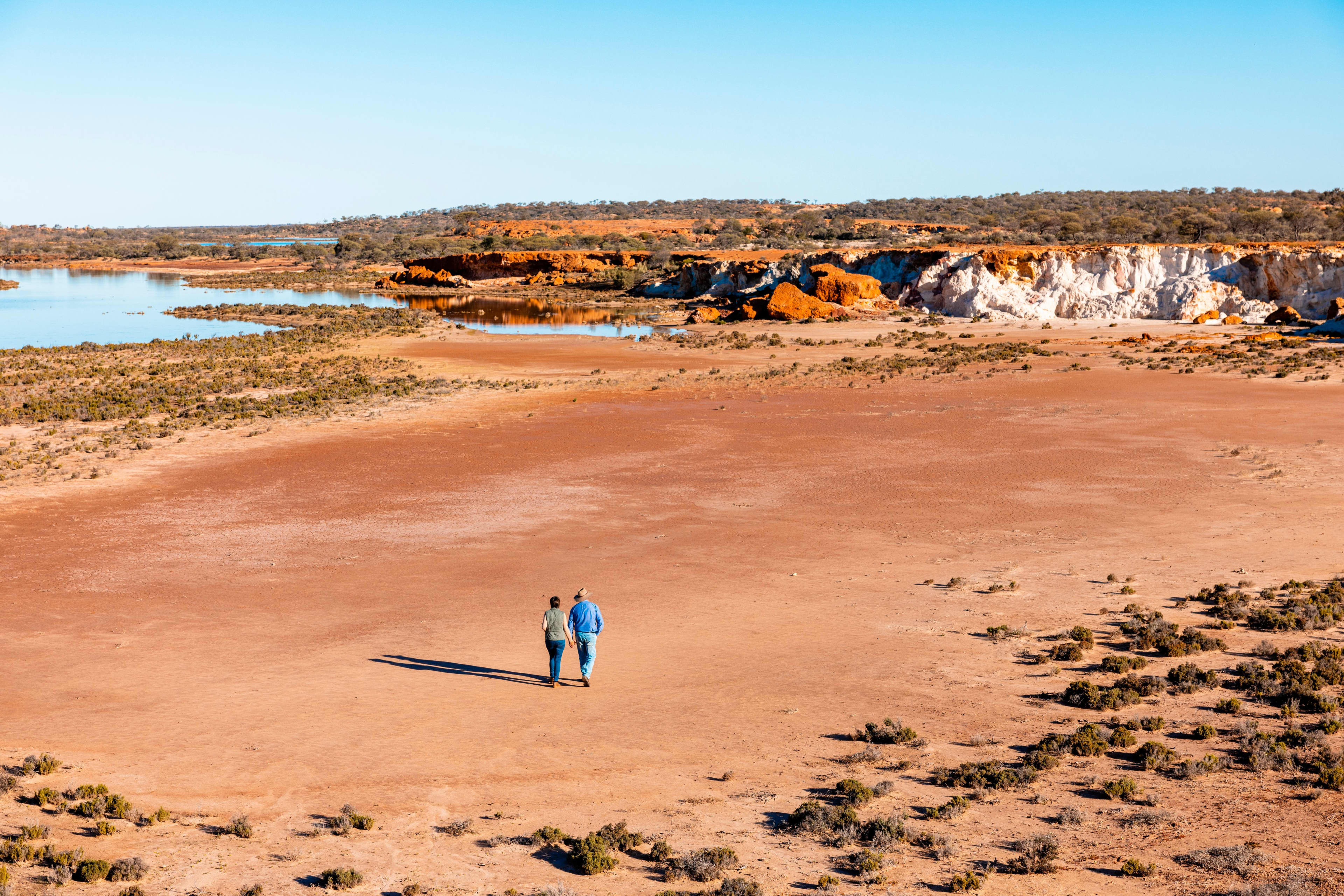 An aerial, wide shot captures a couple walking hand-in-hand across a expansive, flat plain of reddish-brown earth and sand. The couple, consisting of two individuals dressed in blue jeans, are walking away from the viewer toward a bodies of water in the background. Their long shadows stretch out behind them to the left.
