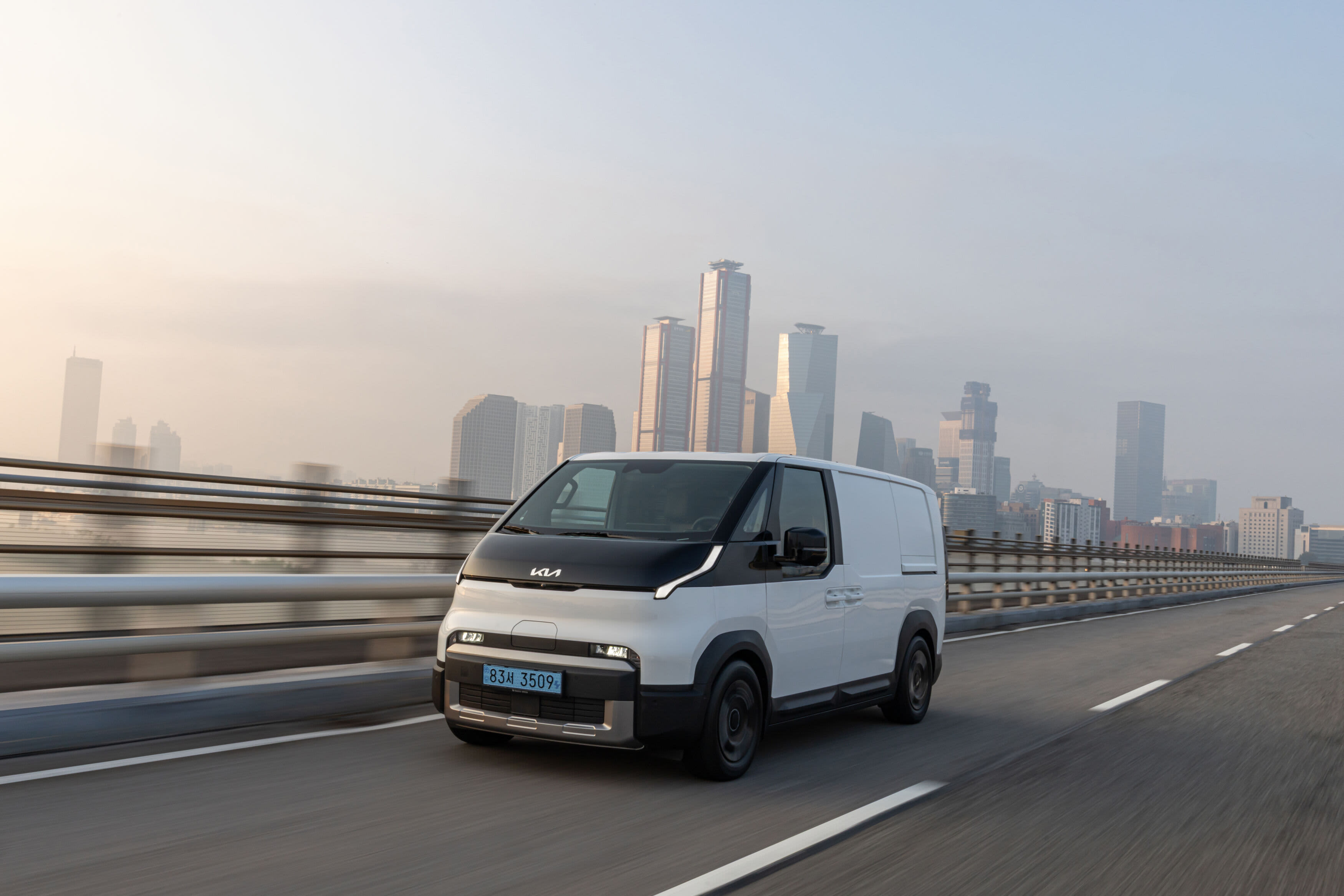 A white Kia PV5 Cargo EV van driving over a bridge near a city