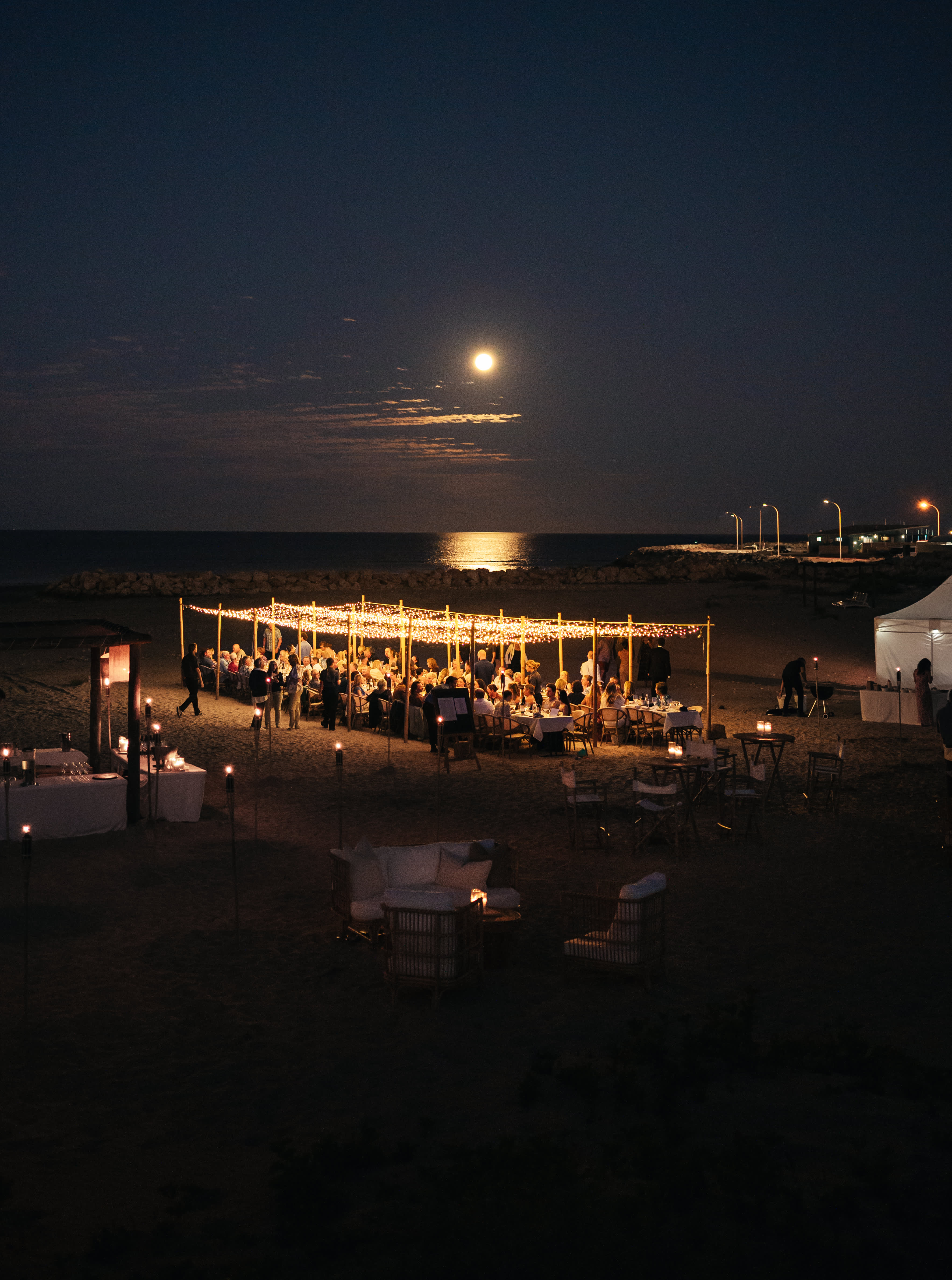 An elevated nighttime view shows a large outdoor dinner gathering on a beach under a full moon. Guests sit at tables beneath a wide wooden canopy illuminated by warm, glowing string lights, while the moon casts a bright reflection across the dark ocean water in the background.