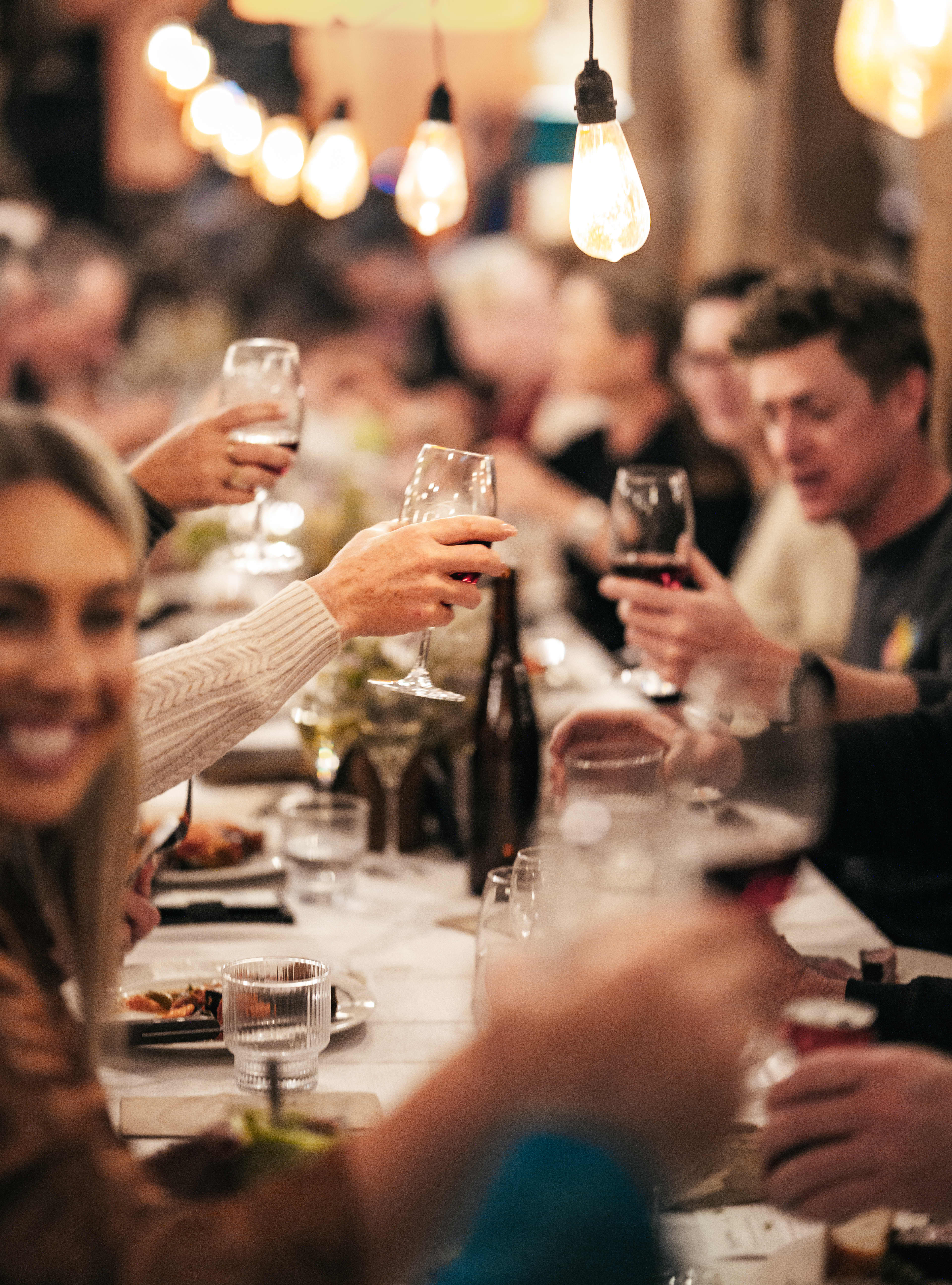 close-up, shallow-depth-of-field view shows guests raising their wine glasses in a toast along a crowded dinner table. Warm, glowing Edison bulb string lights hang directly above, casting a soft blur over the smiling people laughing and interacting in the background.