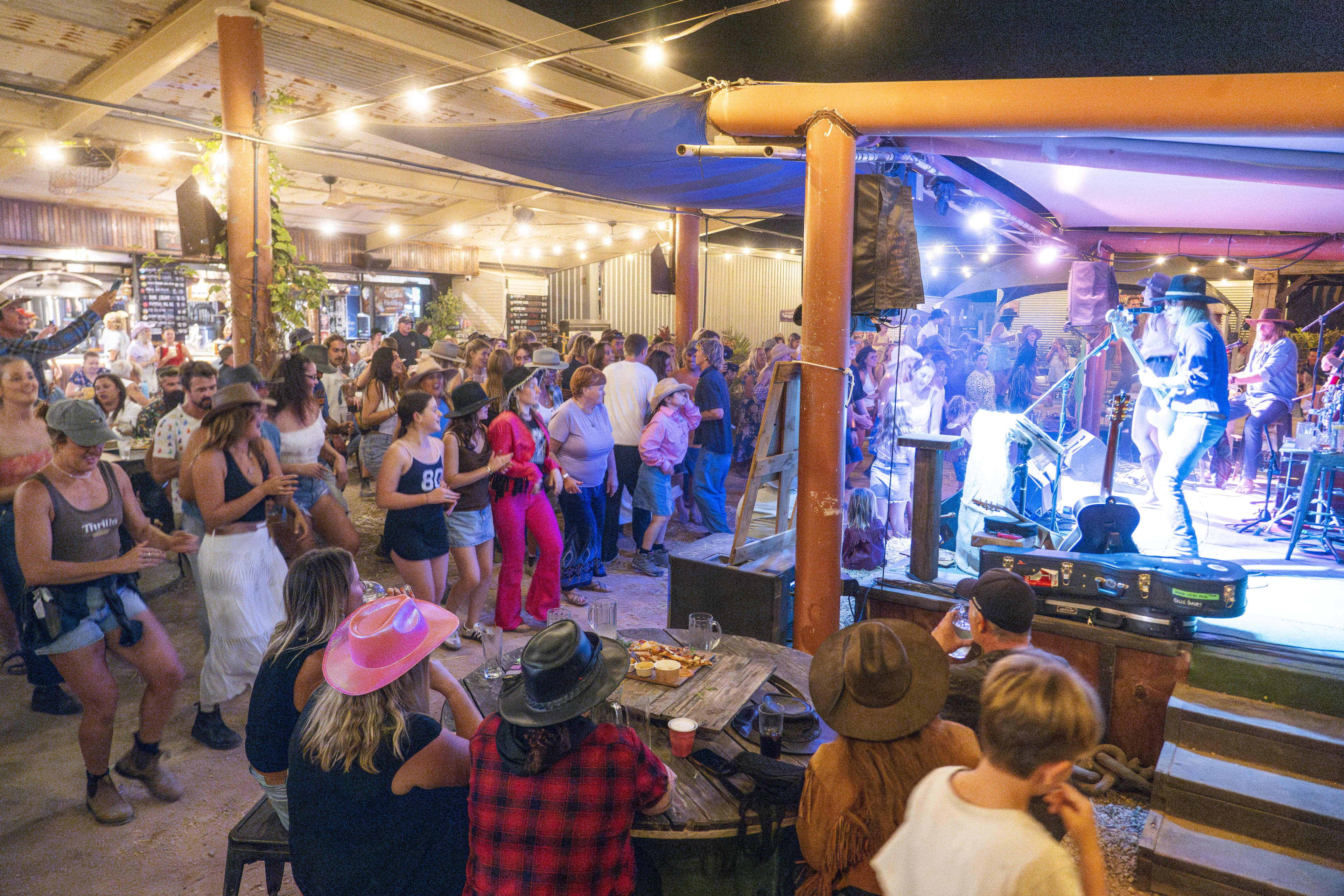 A lively nighttime view shows a crowded outdoor venue where people are dancing and listening to a live band. Musicians perform on a stage illuminated by bright blue lights on the right, while a large crowd, many wearing cowboy hats, dances in an open area lit by overhead string lights.
