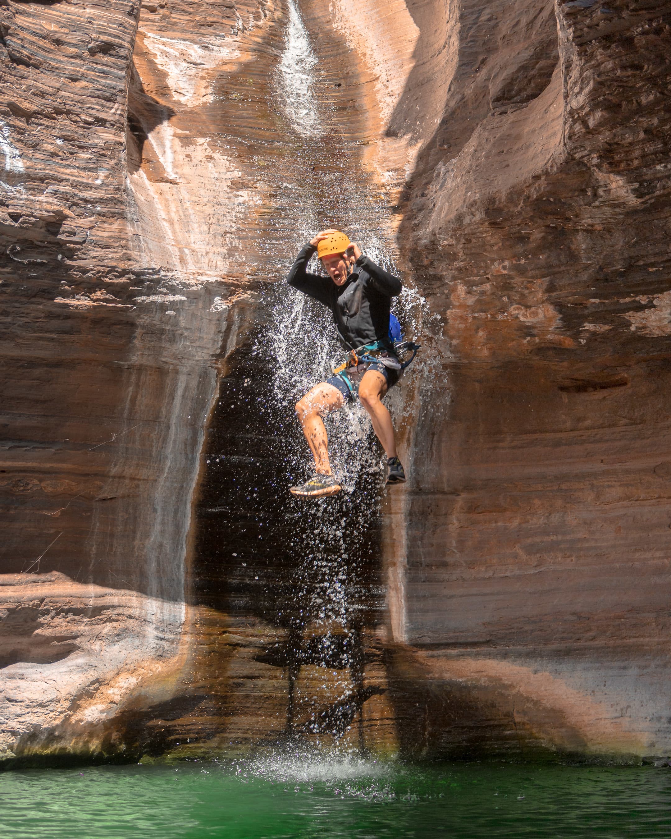 A person wearing a helmet and climbing harness jumps through a small waterfall into a green natural pool surrounded by towering, layered red rock canyon walls.