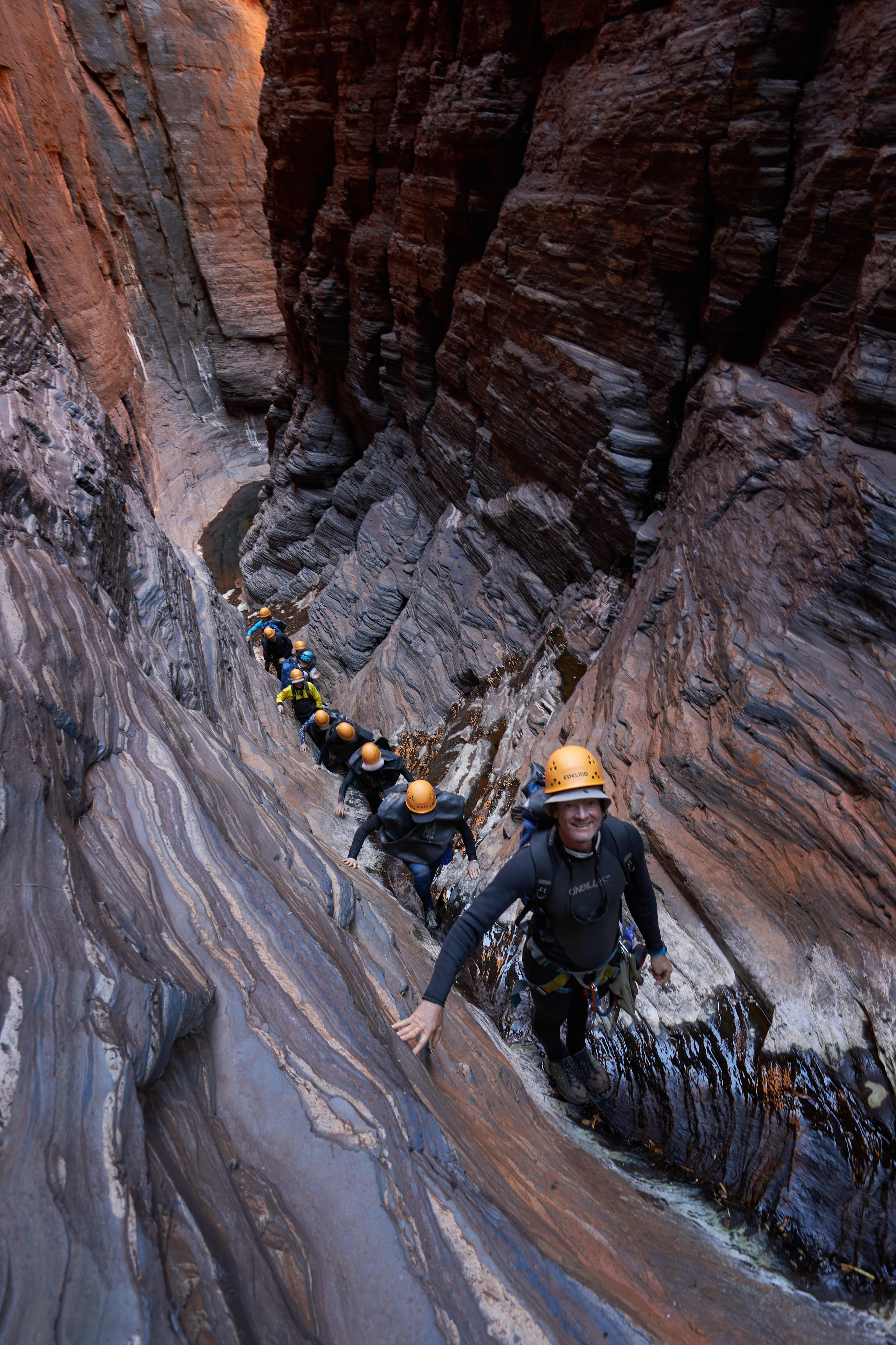 A single-file line of canyoneers wearing yellow helmets and black wetsuits carefully navigates a steep, narrow, and wet rock chute deep inside a towering, layered red stone gorge.