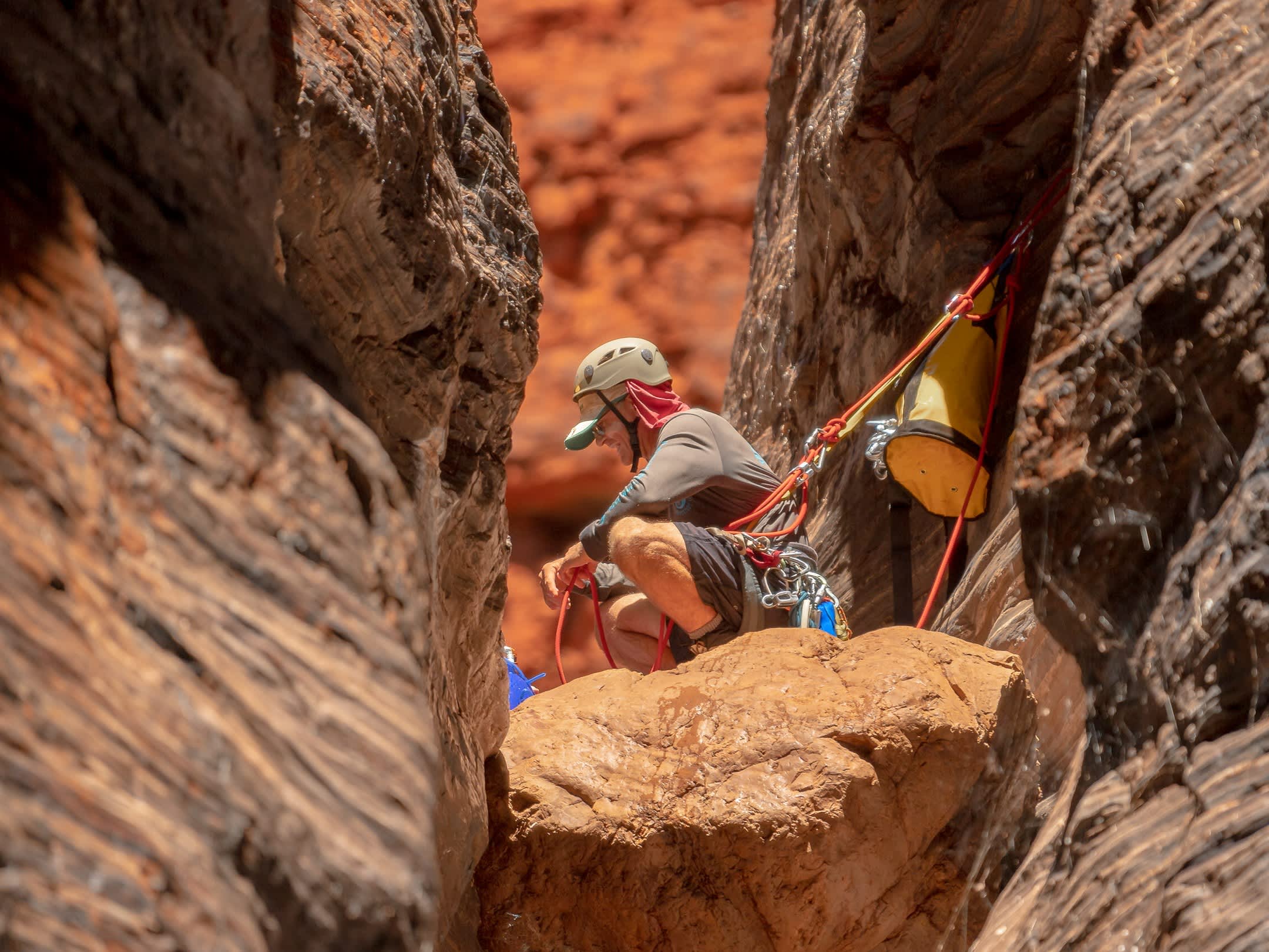 A close-up view of a canyoneer wearing a helmet and safety harness crouching on a large boulder wedged securely within a tight, sun-drenched red rock canyon crevice.