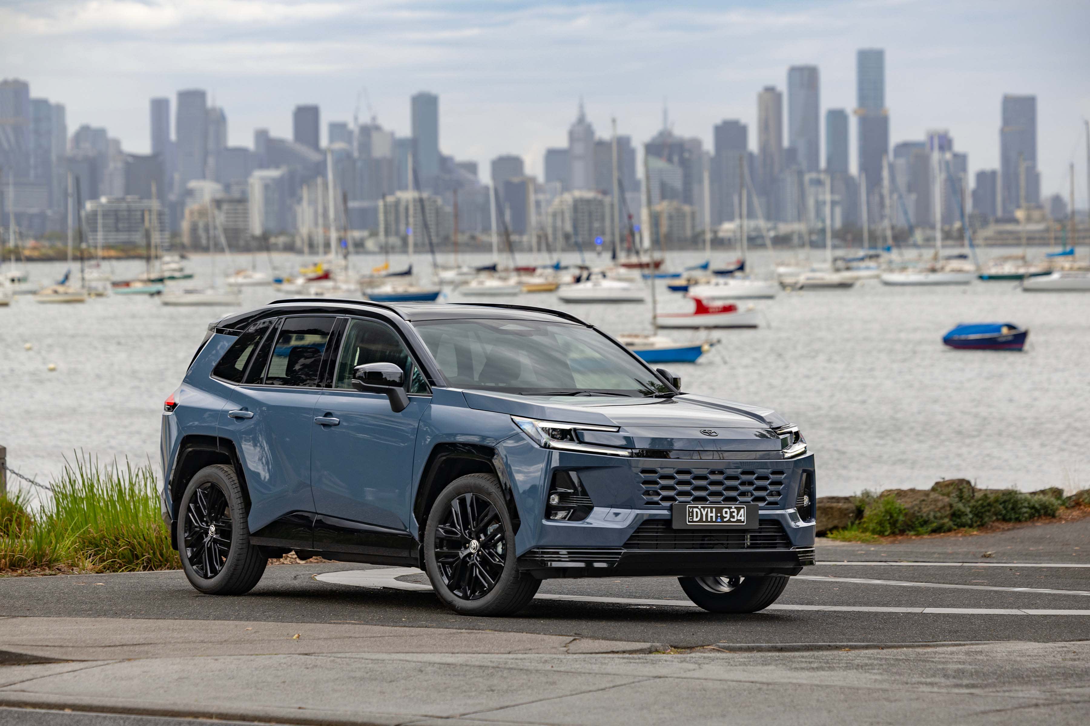 A blue Toyota RAV4 HEV parked near a waterway with city buildings behind