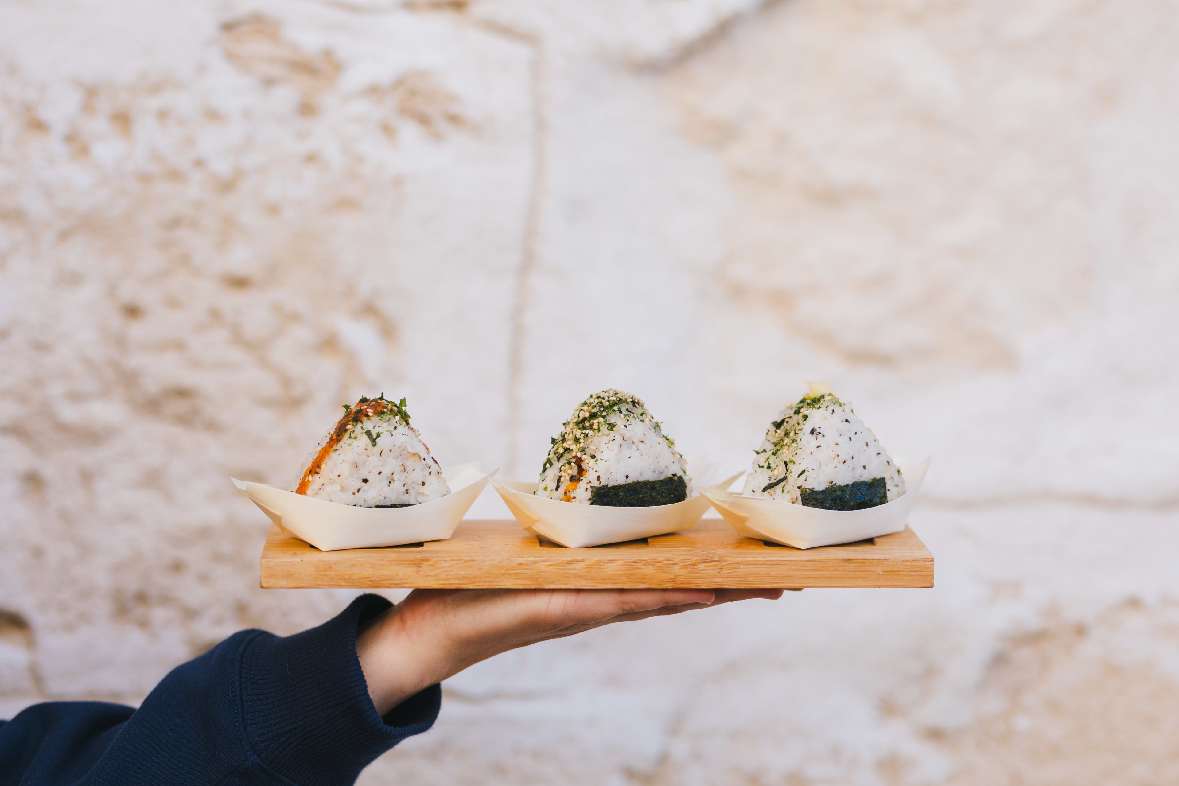 A hand holds a narrow wooden tray displaying three triangular rice balls, known as onigiri, each served in a small paper boat. The onigiri are wrapped with nori seaweed at the base and garnished with different toppings, including green seaweed flakes and a drizzle of orange sauce.