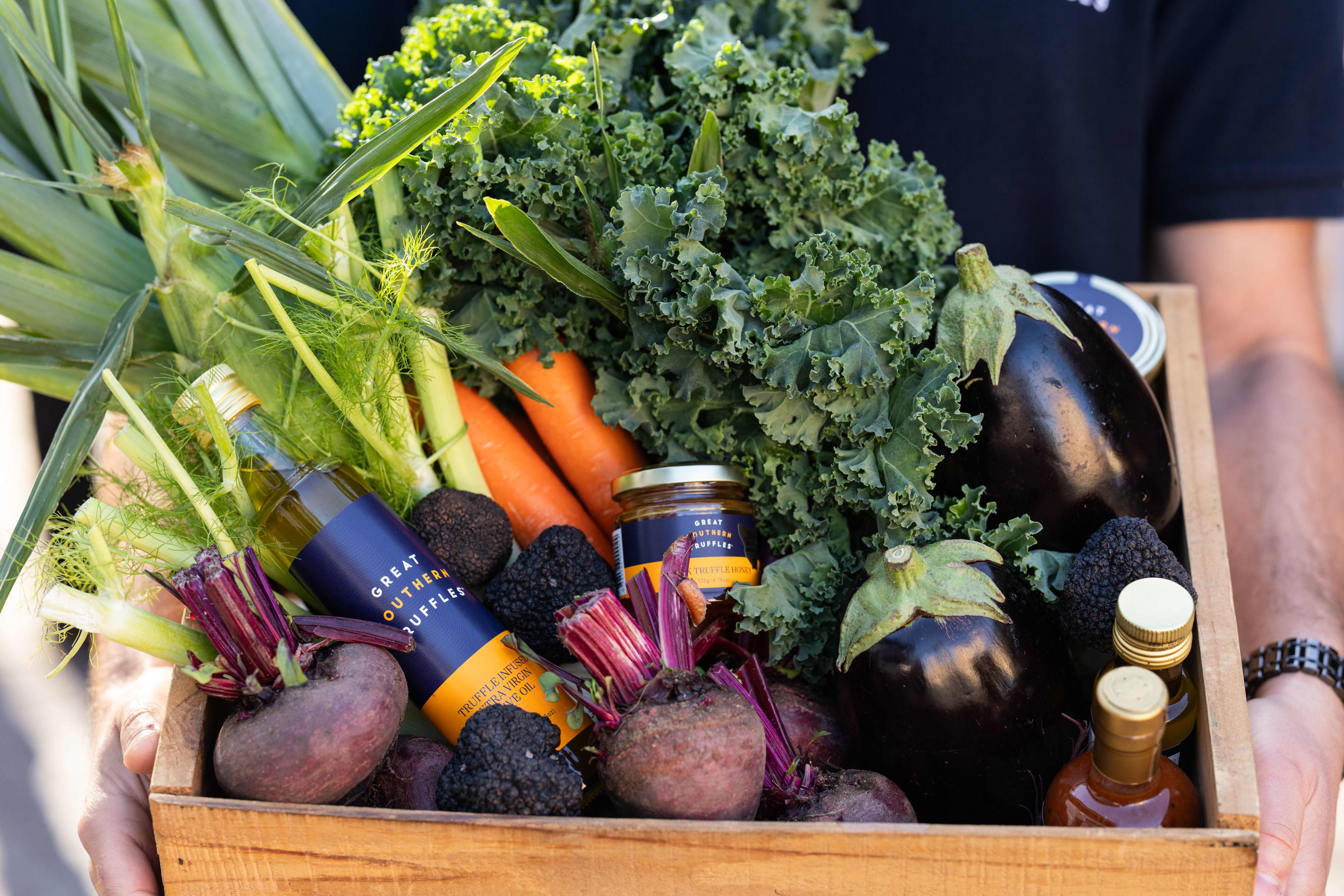 A person holds a wooden crate packed with a variety of fresh produce and gourmet items from "Great Southern Truffles." Inside the crate are leafy green kale, bright orange carrots, two large glossy eggplants, several beetroots with their stems attached, fresh fennel stalks, and several whole black truffles scattered among a bottle of truffle-infused olive oil and a small jar of truffle honey.