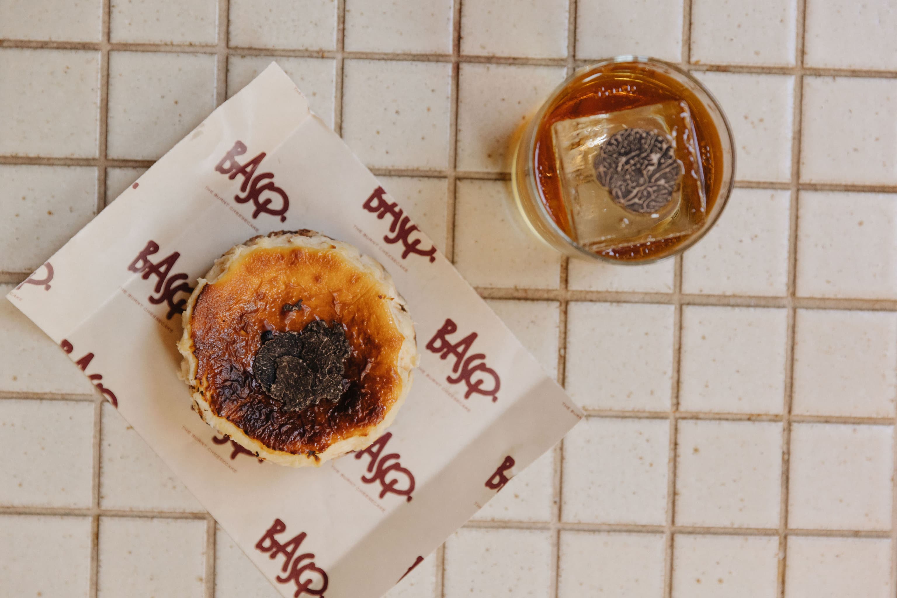 A Basque cheesecake topped with fresh black truffle slices sits on branded paper next to an amber cocktail served over a large ice cube topped with shaved truffle, photographed from above.