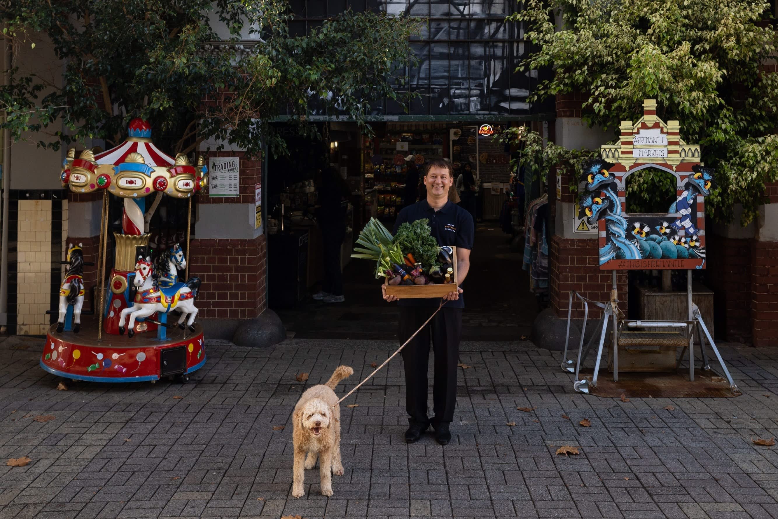 A person stands at the entrance to Fremantle Markets holding a wooden crate filled with fresh vegetables while a curly-haired dog on a lead stands in front. A children's carousel and a decorative market photo frame flank the entrance.