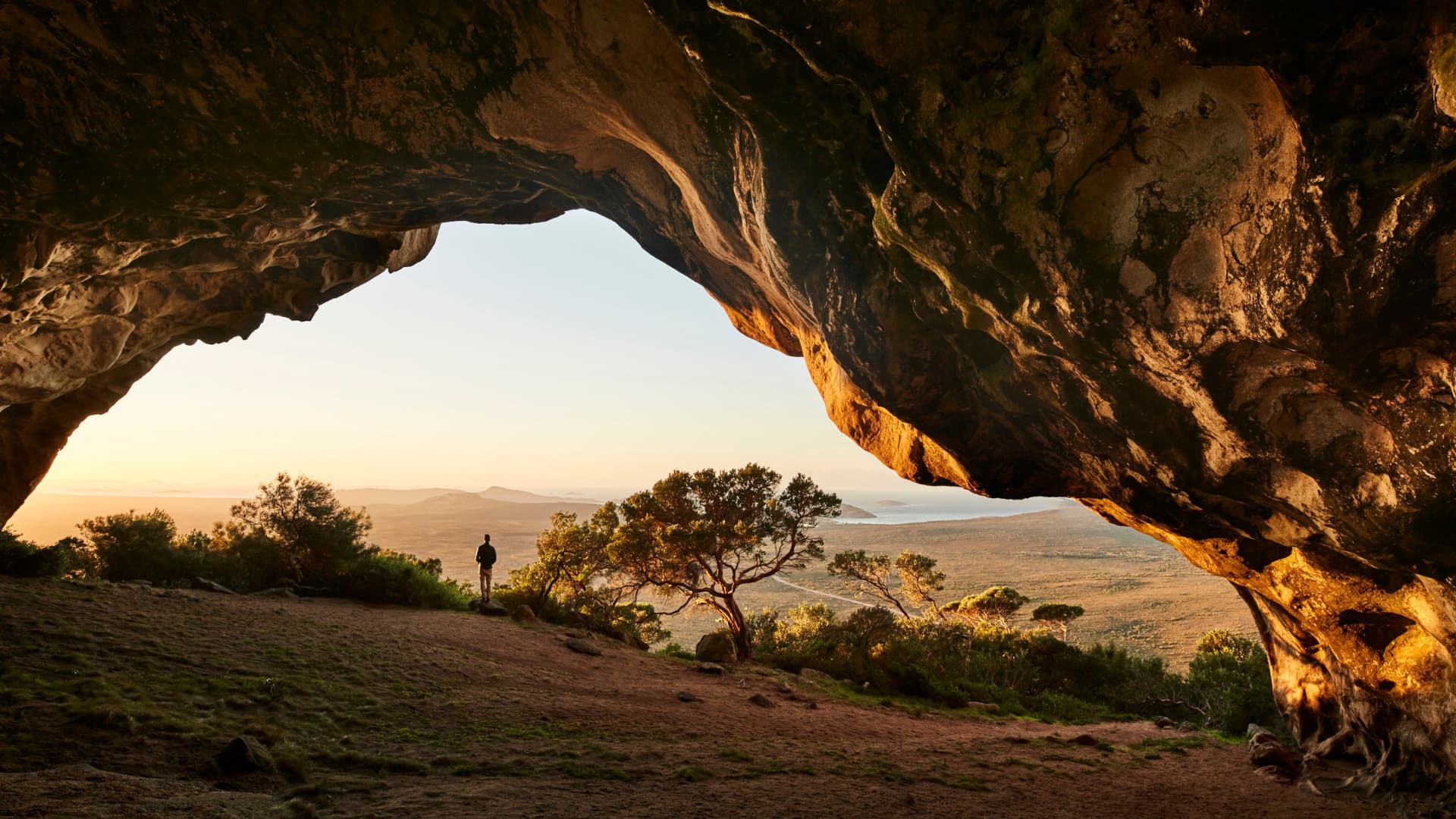 A person stands at the entrance of a massive, textured rock cave, looking out over a vast coastal landscape at sunrise. The dark, curved opening of the cavern frames a sweeping view of low-lying vegetation, a distant coastline, and a calm sea under a soft, golden sky.