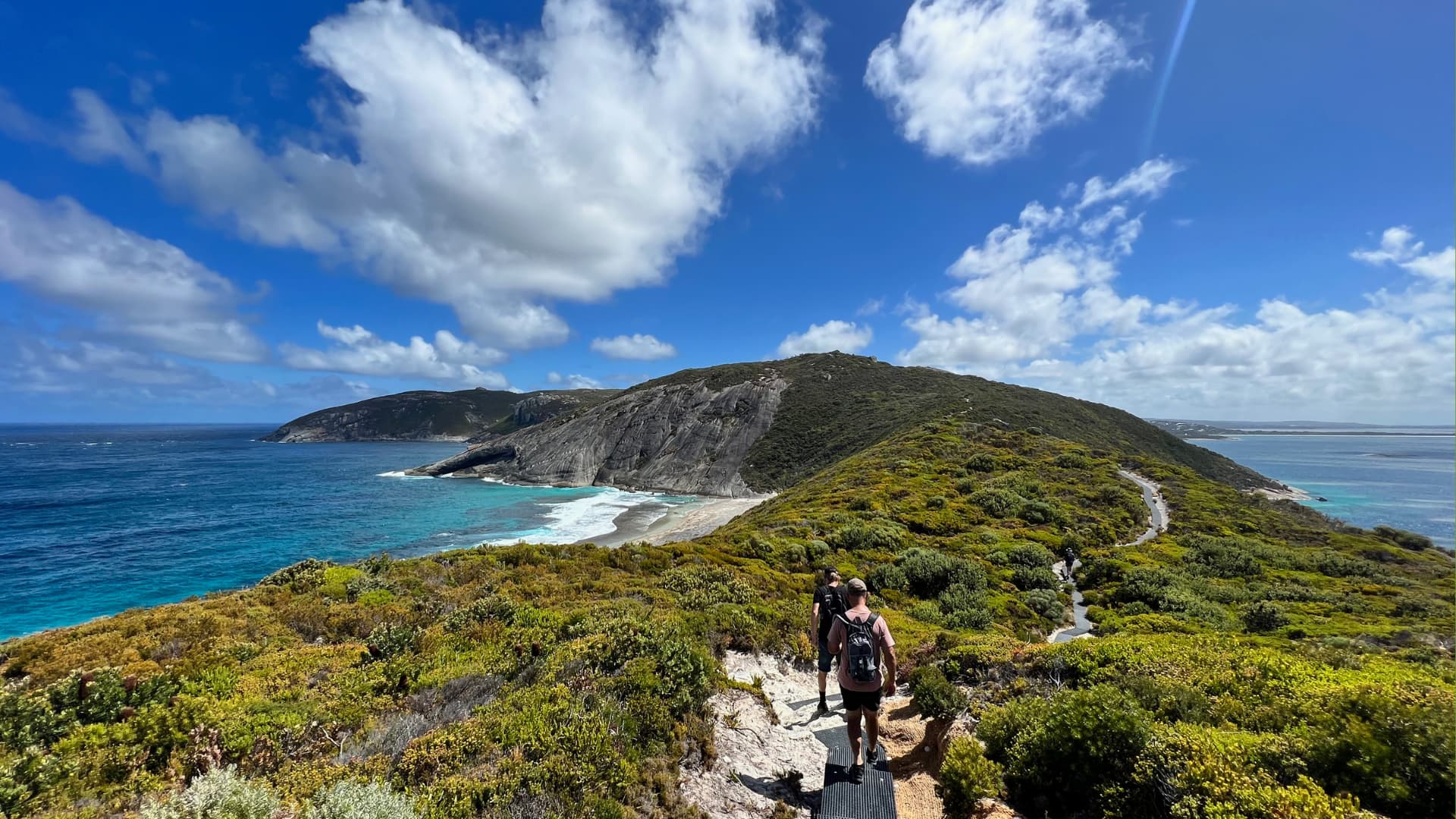 Two hikers walk down a narrow trail along a high coastal ridge covered in green shrubbery. The path overlooks a turquoise ocean bay, a secluded sandy beach, and a massive, exposed grey granite dome hill rising from the peninsula under a bright blue sky with fluffy white clouds.