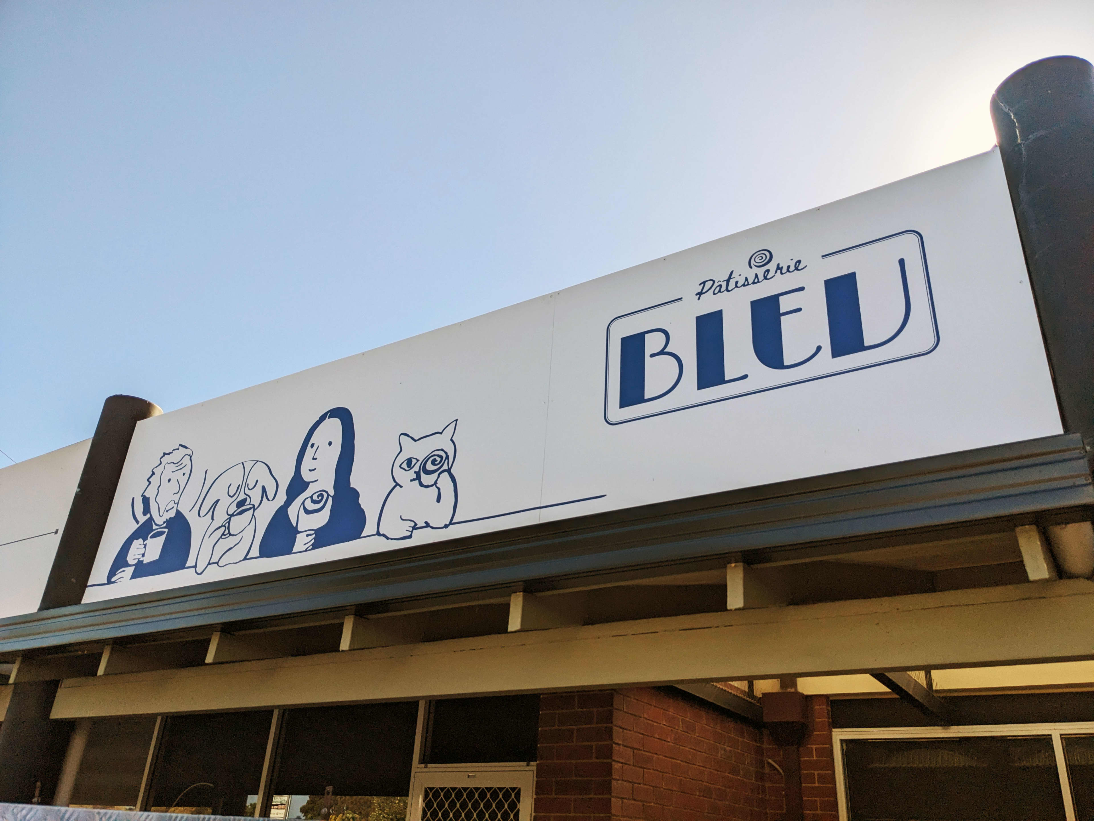 A large white storefront sign reads "Patisserie BLEU" in bold blue lettering inside a thin blue border. To the left of the text, a blue line illustration features a man, a dog, a woman, and a cat, all depicted drinking from mugs or holding treats. The sign is mounted on an outdoor building awning against a clear, bright sky.