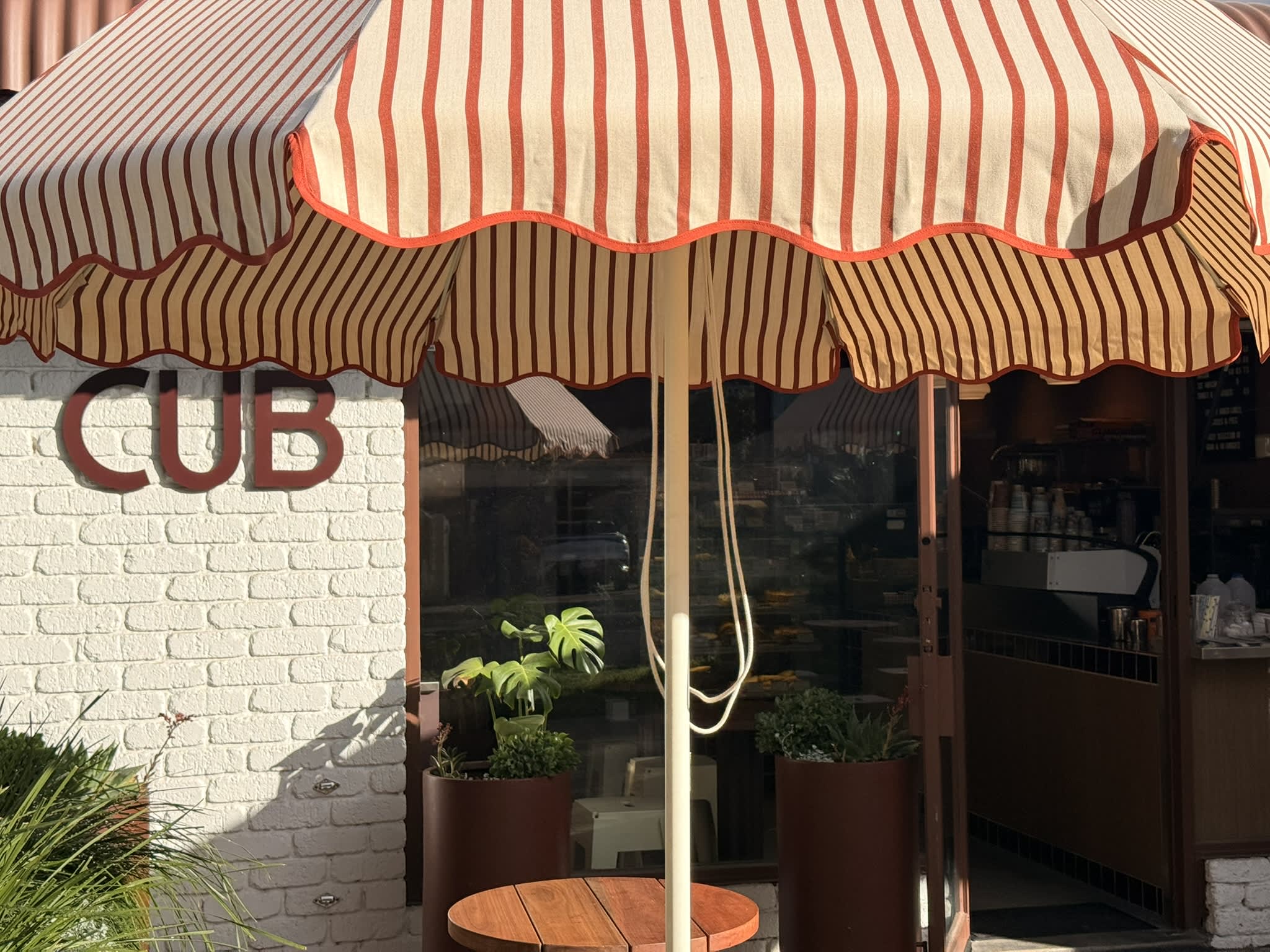 An outdoor cafe storefront named "CUB" features a large red and cream vertically striped umbrella shading a small wooden table. The name "CUB" is mounted in large, dark red block letters on a painted white brick wall to the left. Underneath the umbrella, potted green plants stand near a glass window and door that reveal a glimpse of the cafe's indoor counter and coffee equipment.