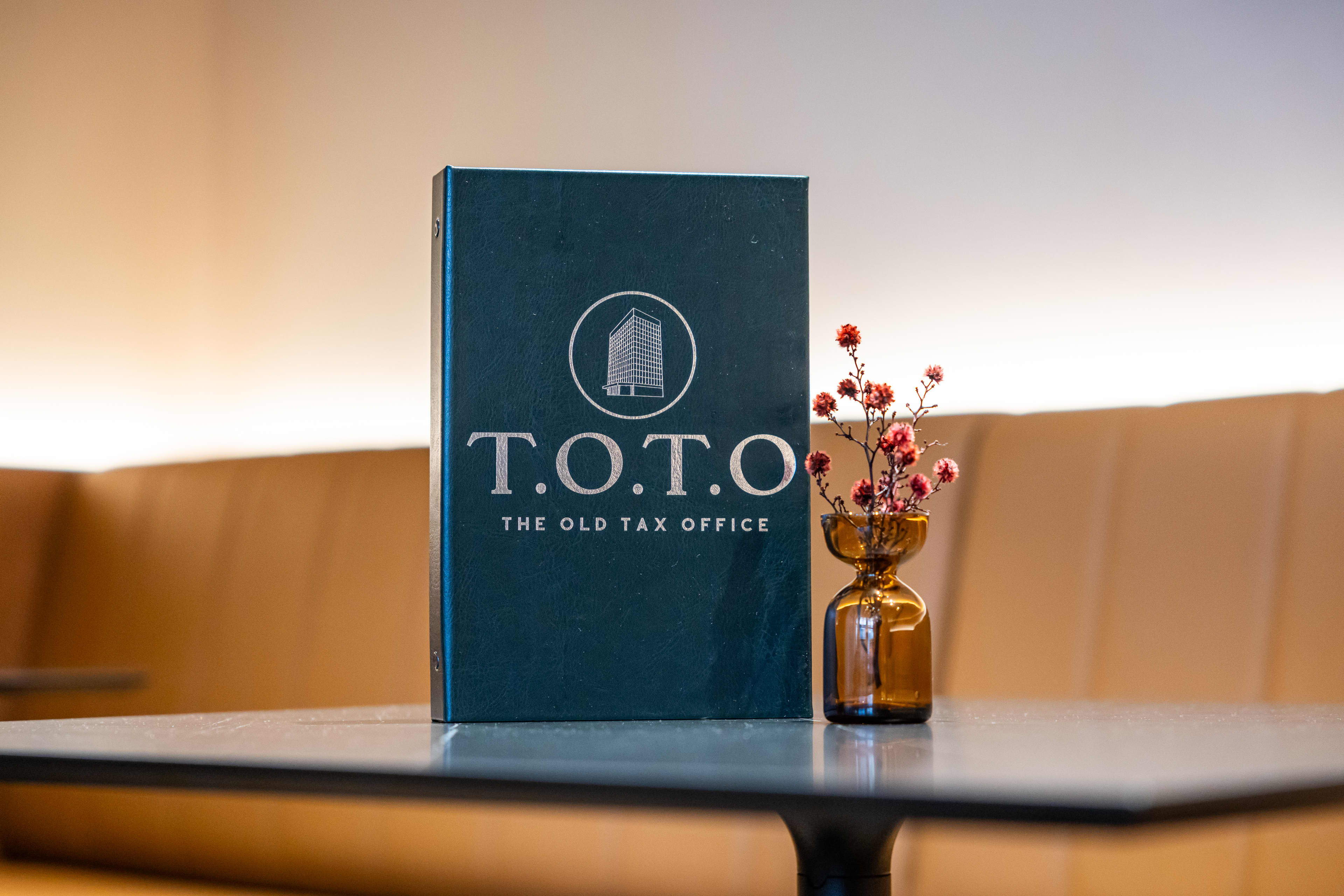 A dark green menu binder stands upright on a dark table next to a small amber glass vase holding tiny pink flowers. The front of the binder features a silver logo showing a building illustration inside a circle, above the text "T.O.T.O" and the subtitle "THE OLD TAX OFFICE."