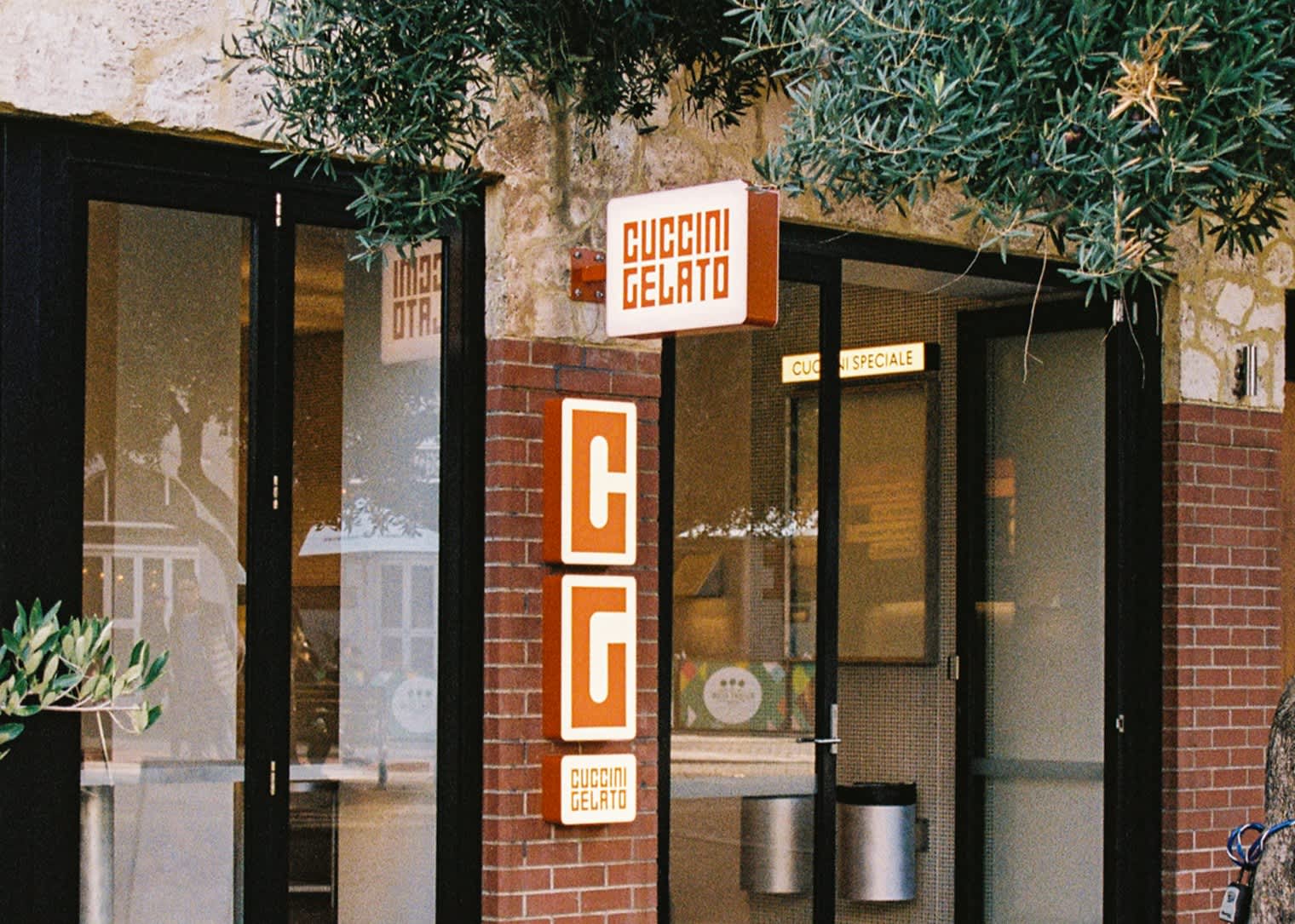 A close-up shot of the exterior storefront of Cuccini Gelato, featuring a rustic stone and red brick facade. A white rectangular sign with orange lettering displays the name "CUCCINI GELATO." Below it, a vertical orange sign showcases a stylized cream-colored "CG" logo. Green olive branches frame the top and left sides of the shop's glass doors and windows.