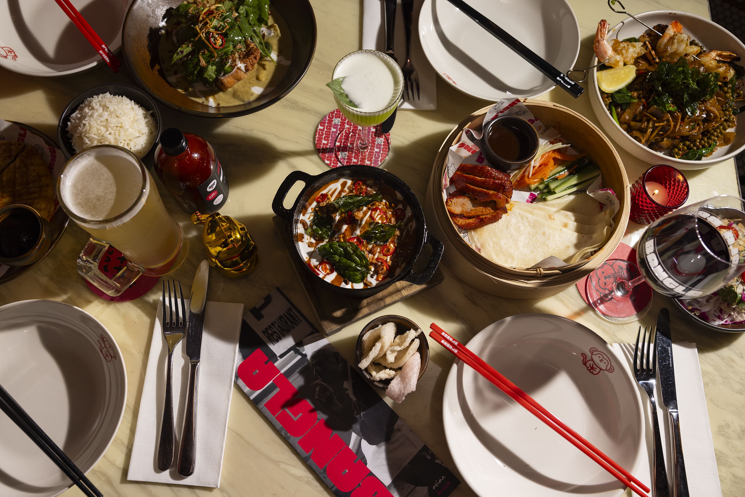 A top-down view of a restaurant table filled with an assortment of Asian dishes and drinks under dramatic lighting. The spread includes a bamboo steamer with roast duck and pancakes, a cast-iron skillet with a red curry dish, stir-fried noodles with prawns, and bowls of rice. Several plates, red chopsticks, a glass of beer, a green cocktail, and a glass of red wine are arranged around a graphic restaurant menu.