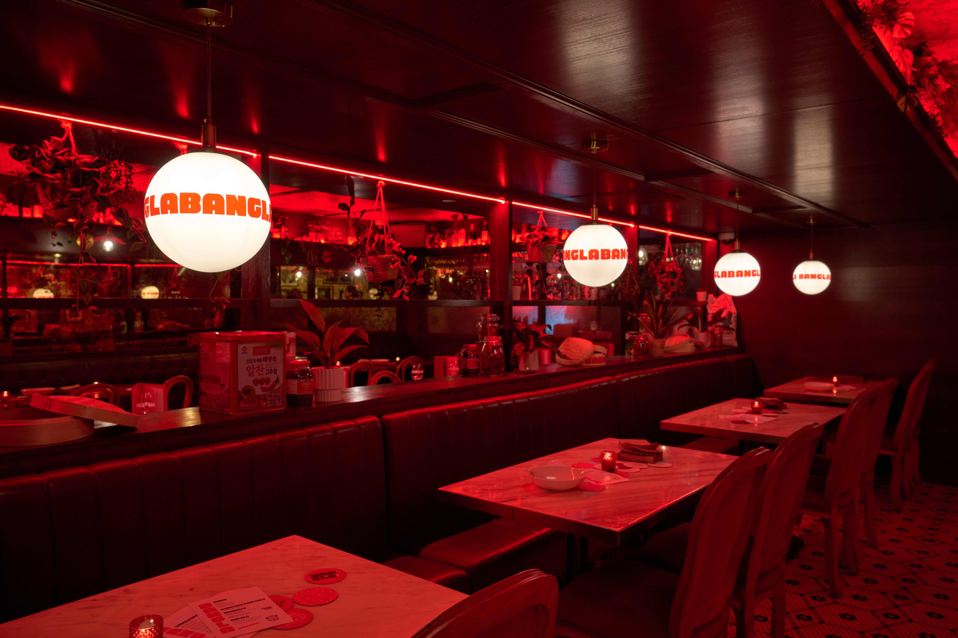 An interior view of a dimly lit restaurant bathed in an intense red neon glow. A row of empty dining tables with marble tops and red chairs lines the right side of the space. Large, spherical white pendant lights hang above the tables, each featuring the bold, black text "LABANGLABANGLABANG" repeated around its circumference.