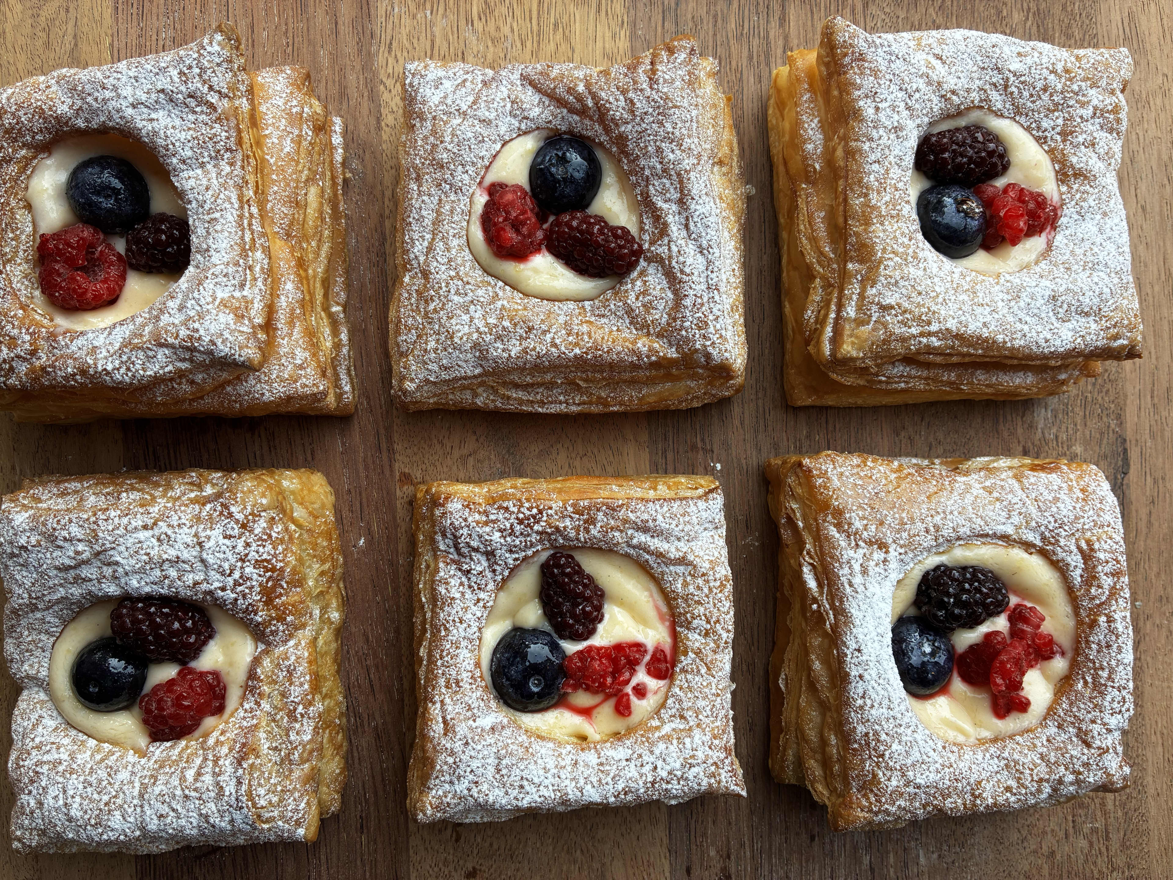 An overhead shot shows six square puff pastry danishes arranged in two rows of three on a wooden surface. Each pastry has a golden-brown, flaky crust dusted with powdered sugar, creating a square border around a center filling of pale yellow custard. Nestled inside the custard of each danish are three fresh berries, including a mix of deep blue blueberries, red raspberries, and dark blackberries.