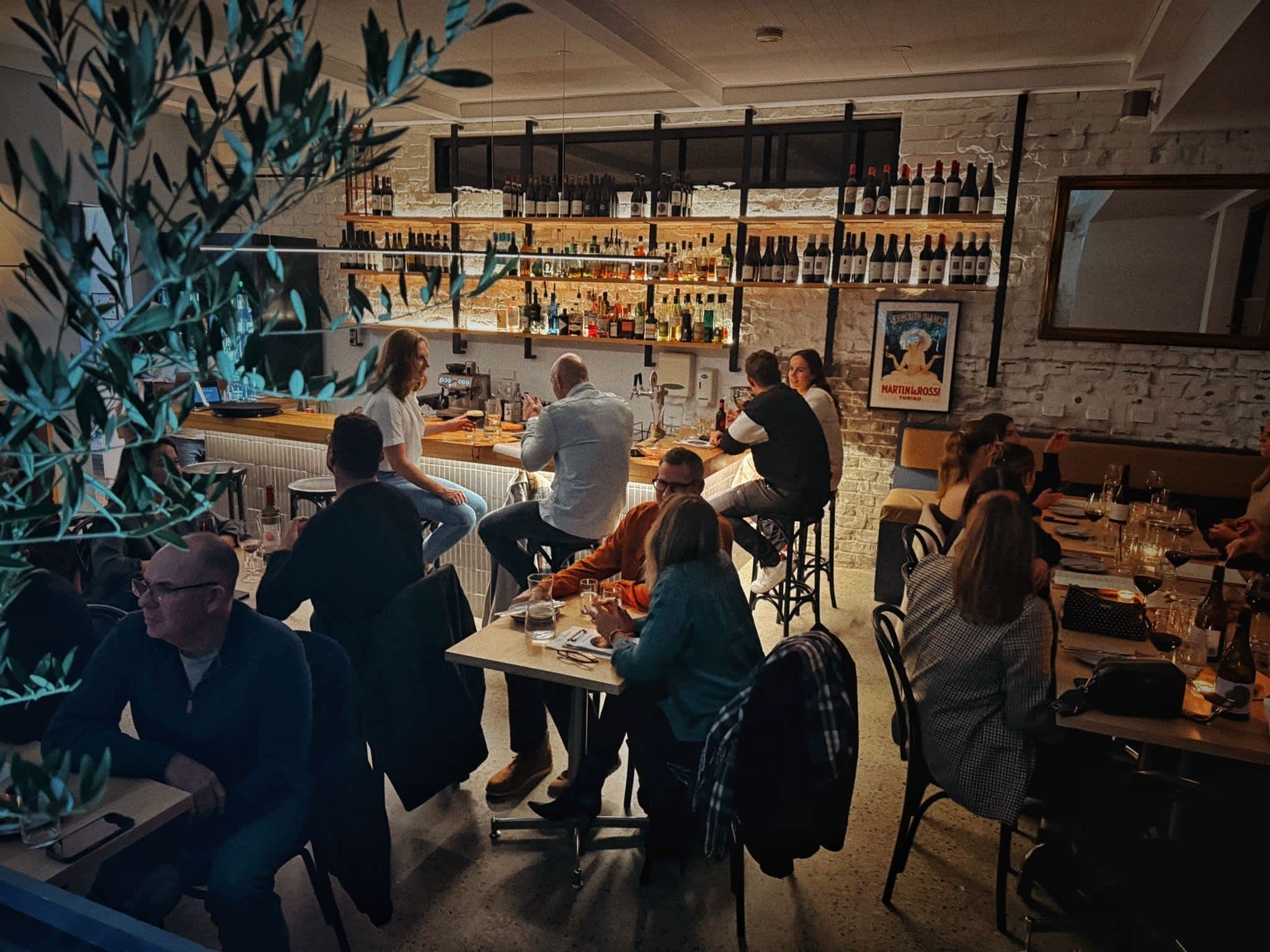 A dimly lit, bustling restaurant interior shows patrons seated at tables and along a wooden bar counter. Multiple rows of wine bottles line open backlit shelves against a white brick wall behind the bar. In the foreground, people sit at small dining tables, while olive tree branches frame the left side of the view.