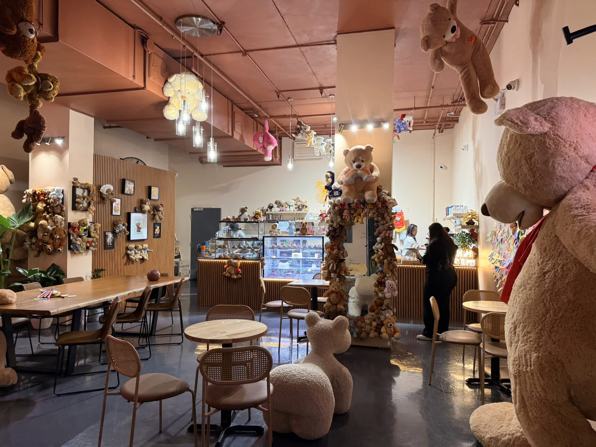 A brightly lit cafe interior features a pink ceiling and is heavily decorated with plush teddy bears of various sizes hanging from above and mounted on walls. Wooden dining tables and modern chairs are spread across the polished gray floor, leading toward a service counter with a pastry display case in the background. A giant teddy bear dominates the right foreground, while a plush bear-shaped chair sits near the centre.