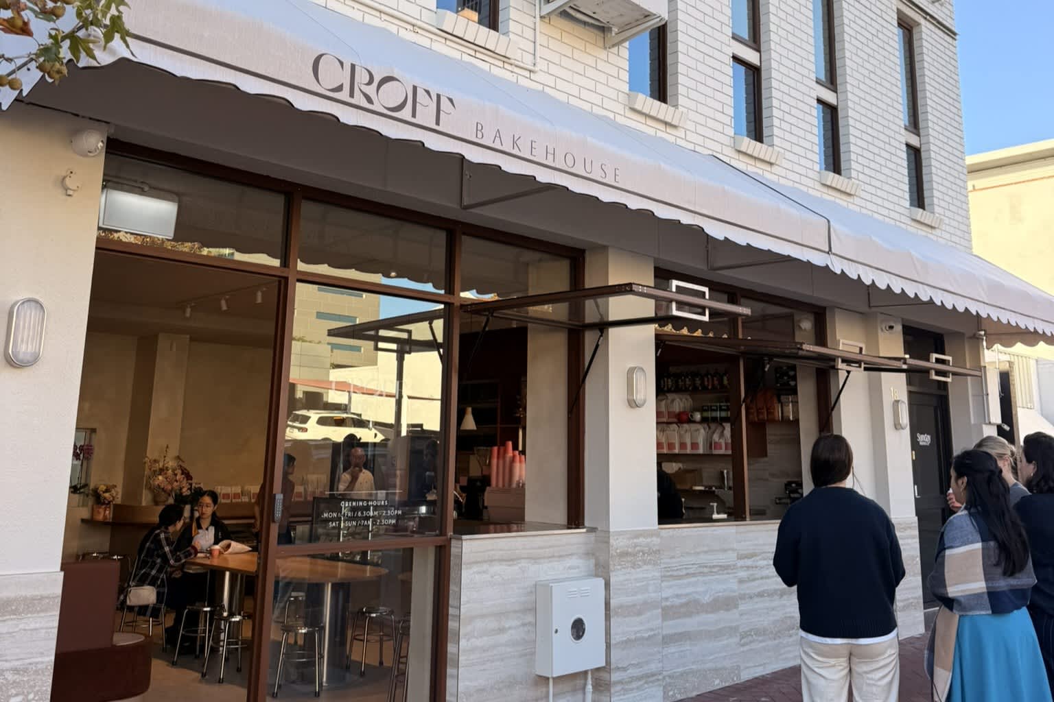 The exterior of a modern bakery named "CROFF BAKEHOUSE" features a white brick building with a large white awning and expansive glass windows. Customers wait in a line along the outdoor brick pavement next to an open service window, while a few patrons are visible seated inside the bright, minimalist dining area.