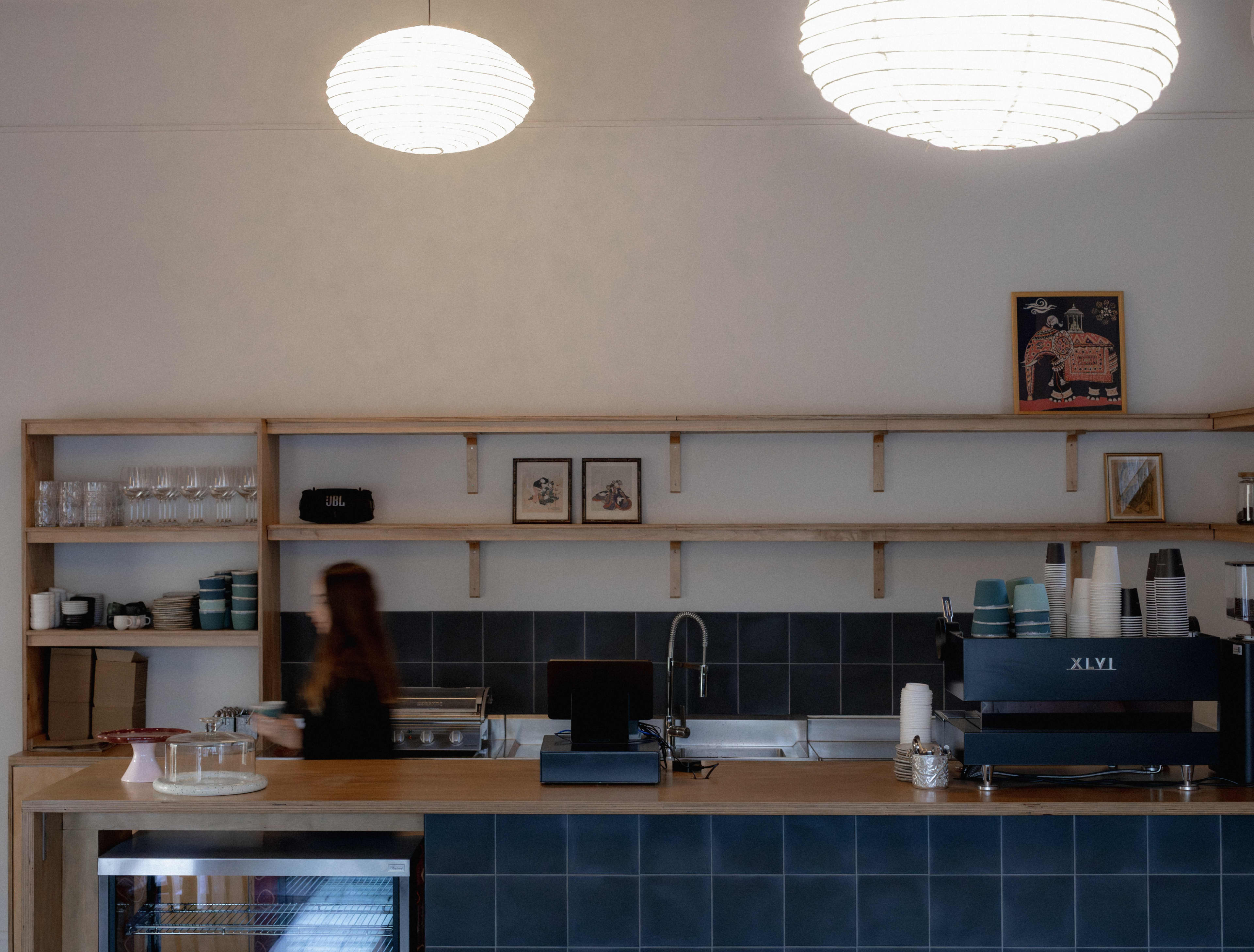 A brightly lit cafe counter featuring a long wooden countertop backed by a dark blue tiled backsplash. Open wooden shelving mounted on the white wall behind the counter holds rows of drinking glasses, stacks of ceramic cups, small framed pictures, and an espresso machine with paper cups stacked on top. Two large, glowing spherical paper lantern lights hang from the ceiling. On the left side of the counter, a person with long hair is captured in motion blur while working behind the bar.