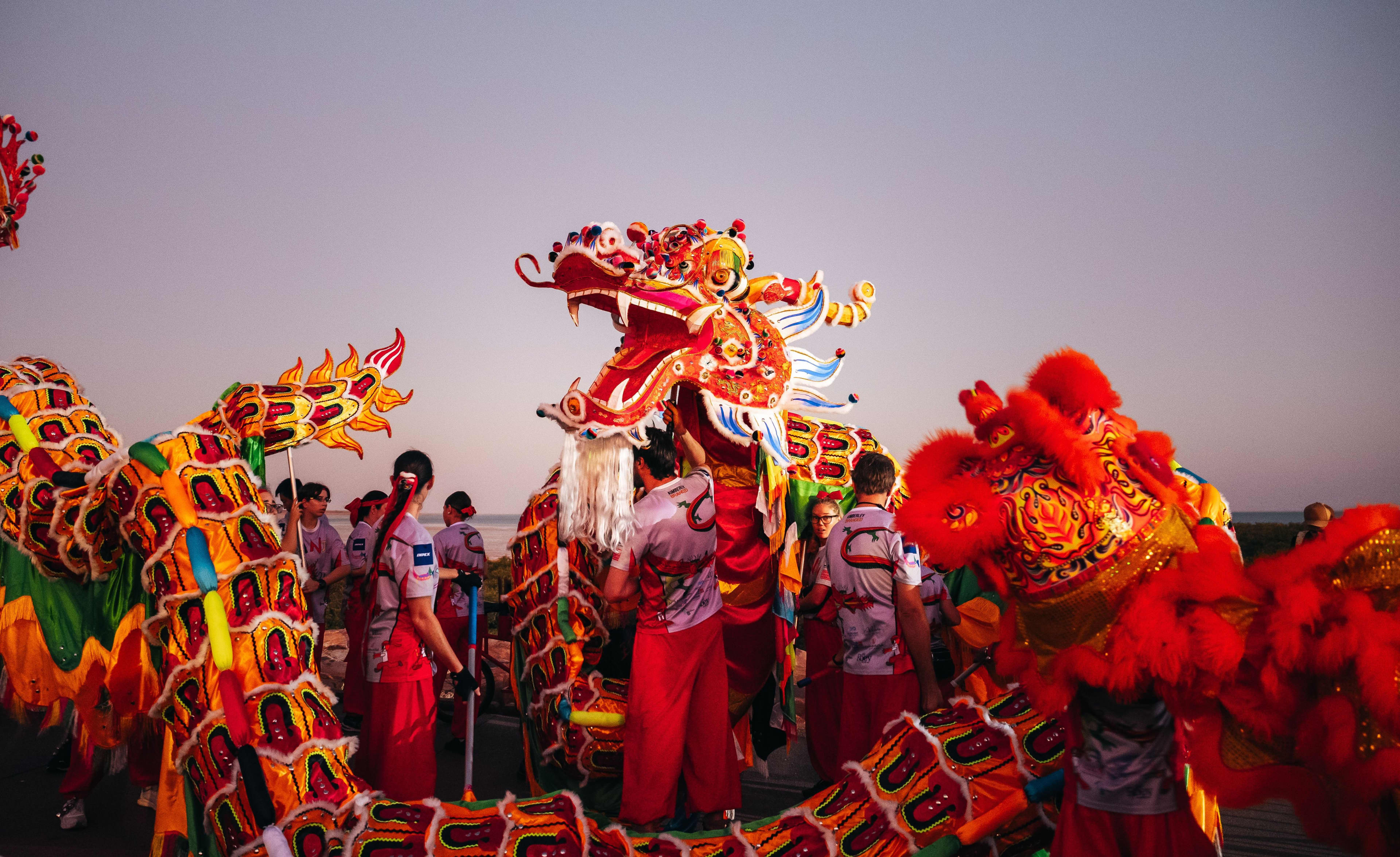 Performers prepare a large, colorful Chinese dragon prop outdoors during twilight. The dragon features an intricate, open-mouthed head decorated in red, orange, and gold, with a long scaly body stretching across the frame. The team wears matching gray t-shirts with a stylized dragon graphic and bright red pants against a soft, gradient sky near the coast.
