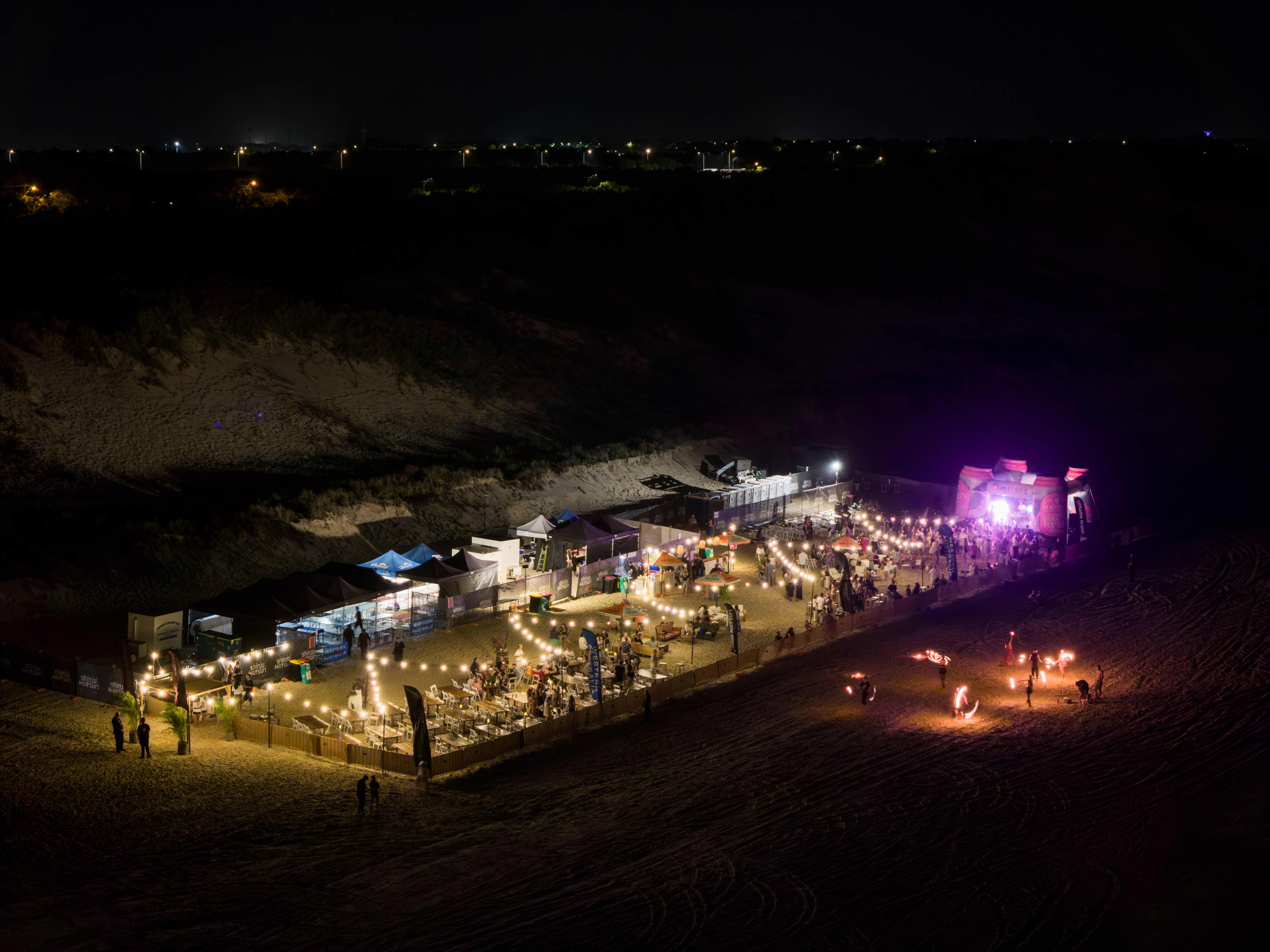 An elevated nighttime view of a bustling beach festival market enclosed on the sand. The market area is brightly illuminated by warm string lights and features rows of white and blue canopy tents, dining tables, and a main stage glowing with purple light at one end. Outside the fence on the dark open beach, fire twirlers perform with glowing flames while city lights flicker in the distant background.