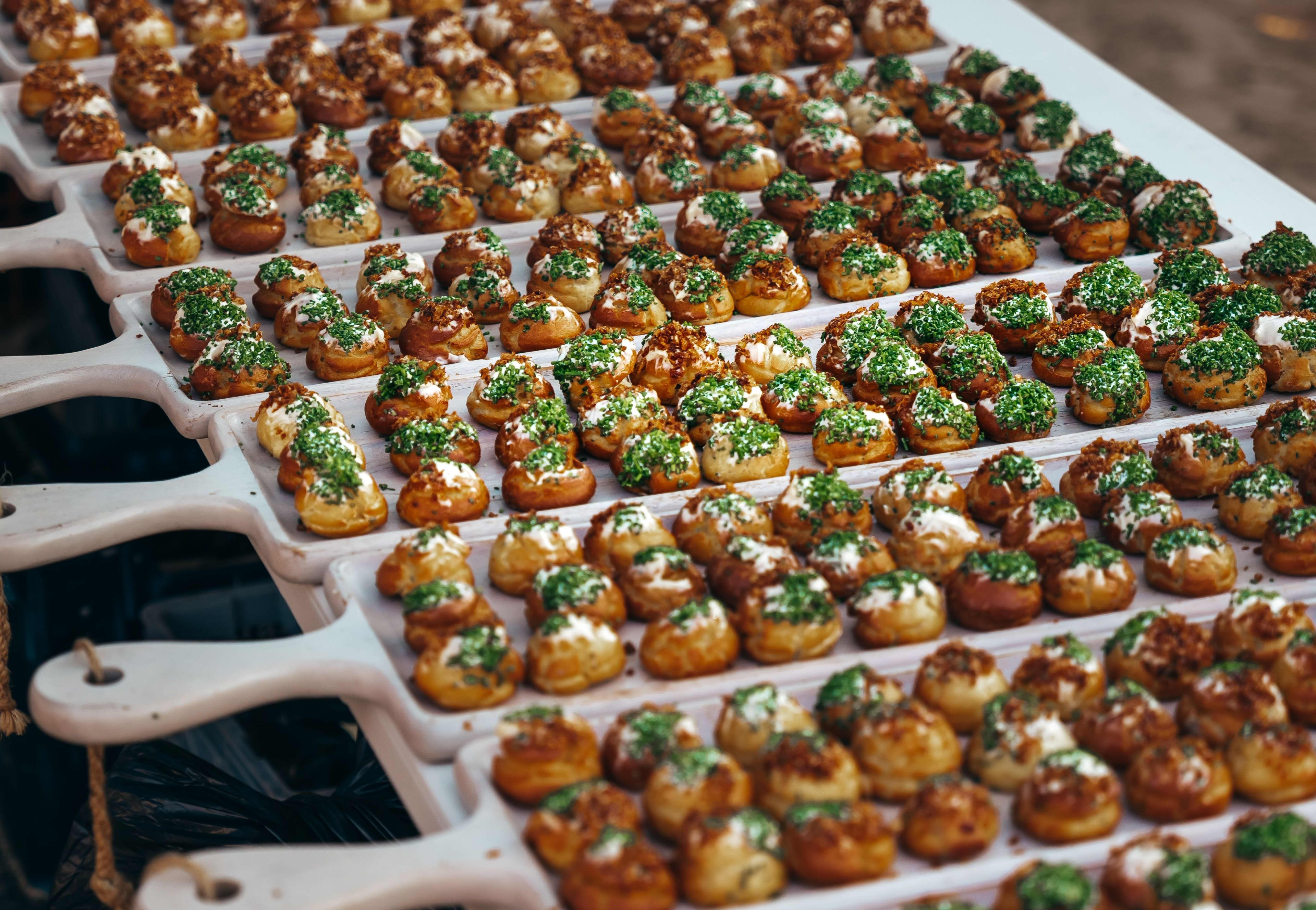 Rows of white wooden serving boards arranged on a table, filled with dozens of small, round, golden-brown bite-sized pastries. The gourmet appetizers feature different toppings, including some covered in fresh chopped green herbs, some dolloped with white cream, and others sprinkled with crispy brown bits.