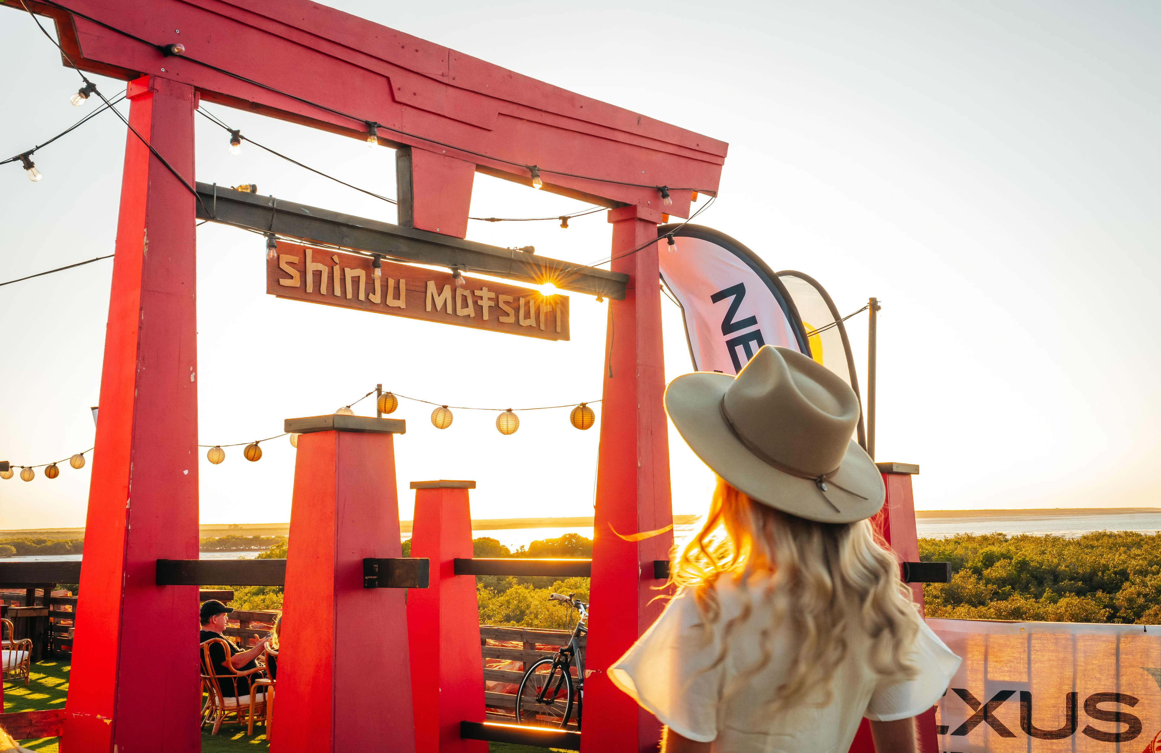 A woman with wavy blonde hair wearing a wide-brimmed fedora hat looks toward a large red torii gate during a golden sunset. A wooden sign hanging from the gate reads "Shinju Matsuri," illuminated by the warm, direct sunlight. String lights and round lanterns hang across the structure, overlooking a landscape of coastal mangroves and a distant body of water under a clear, bright sky.