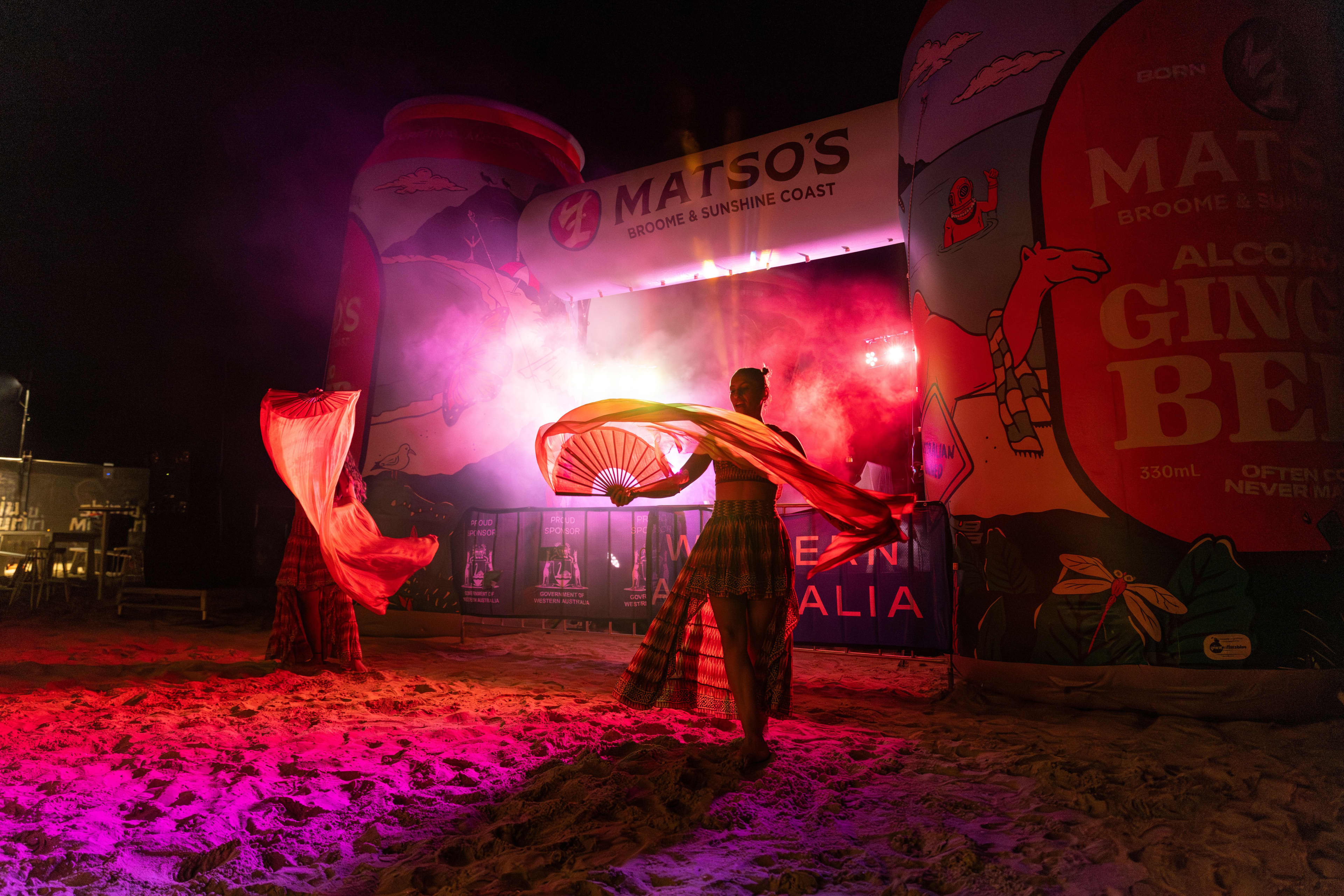 Two performers dance with large, flowing red fans on a sandy floor at night, illuminated by dramatic pink and red stage lighting. A smoke effect swirls behind them under an archway bearing the brand name "Matso's Broome & Sunshine Coast," flanked by massive inflatable cans, including one prominently displaying "Ginger Beer."