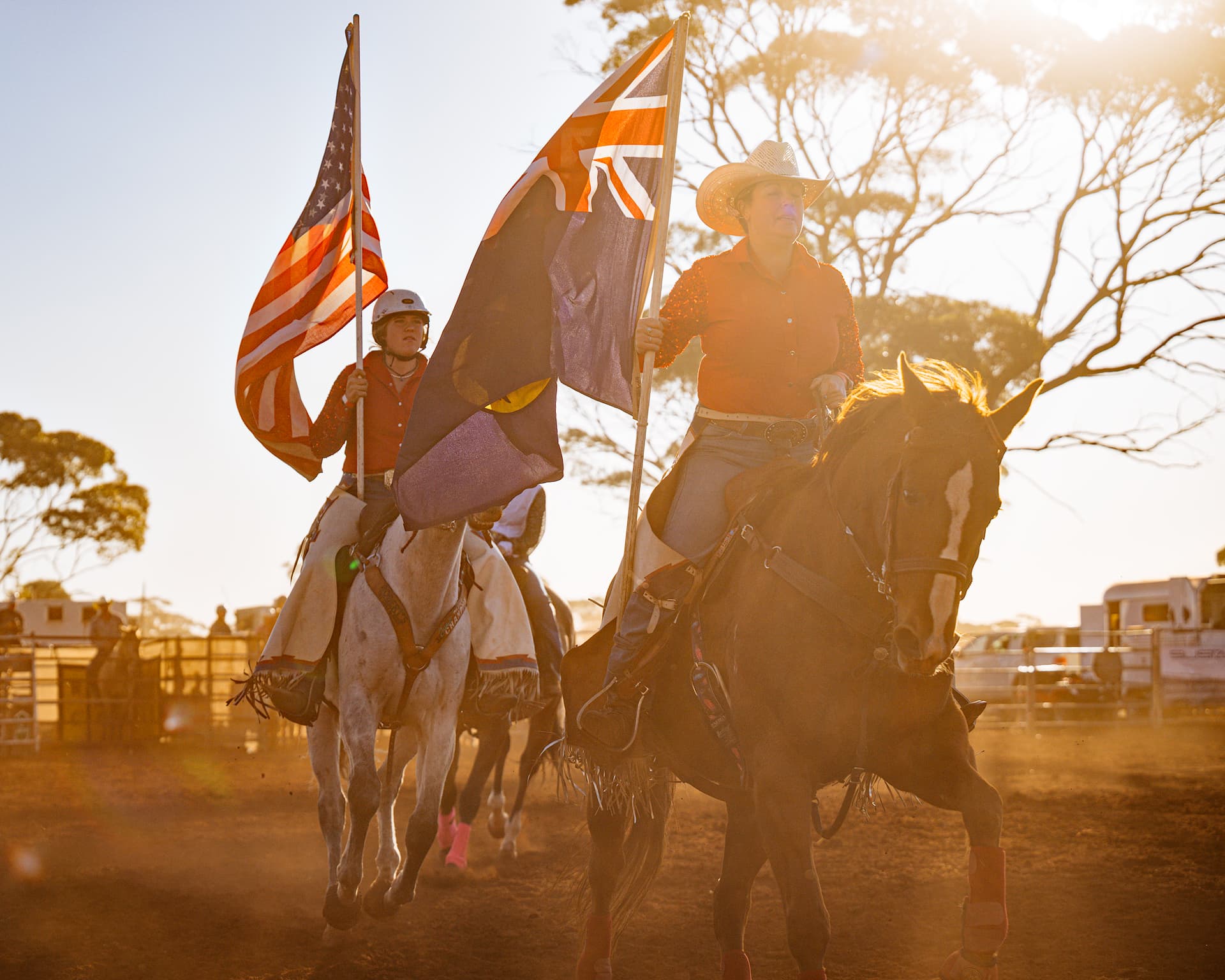 Two riders on horseback carry flags in a dusty arena during golden hour. On the right, a woman in a cowboy hat and orange shirt rides a brown horse, holding the Australian flag. Behind her to the left, another rider on a white horse carries the American flag. The bright evening sun creates a strong lens flare, illuminating trees and trailers in the background.