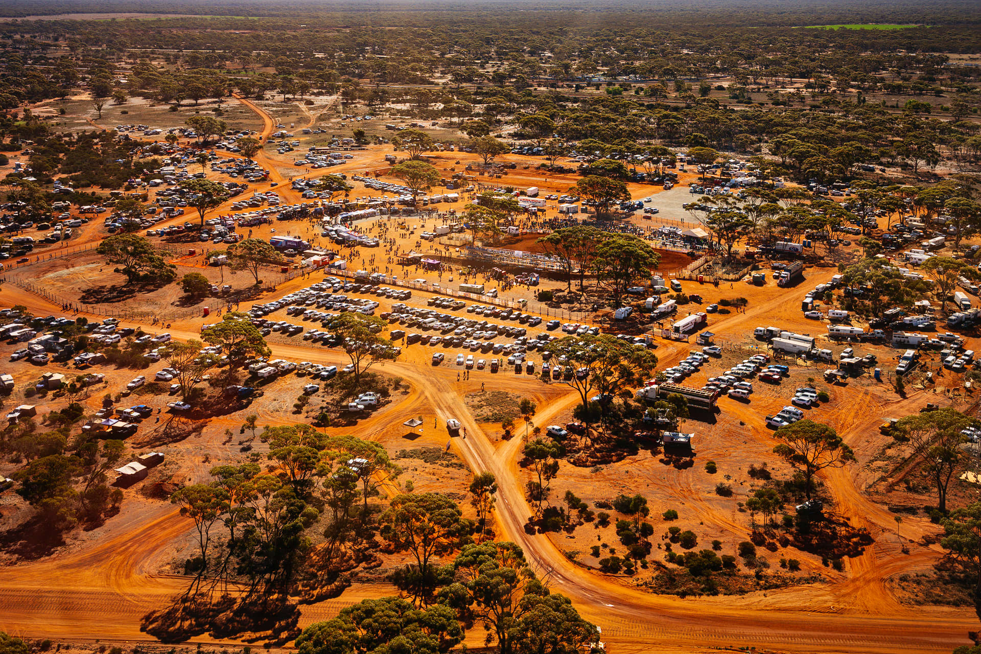 An aerial view captures a large event grounds situated in a vast, arid outback landscape filled with sparse green trees and bright orange dirt roads. At the center, hundreds of parked cars and trucks form organized rows, surrounded by temporary campsites, trailers, and event structures scattered throughout the bush.