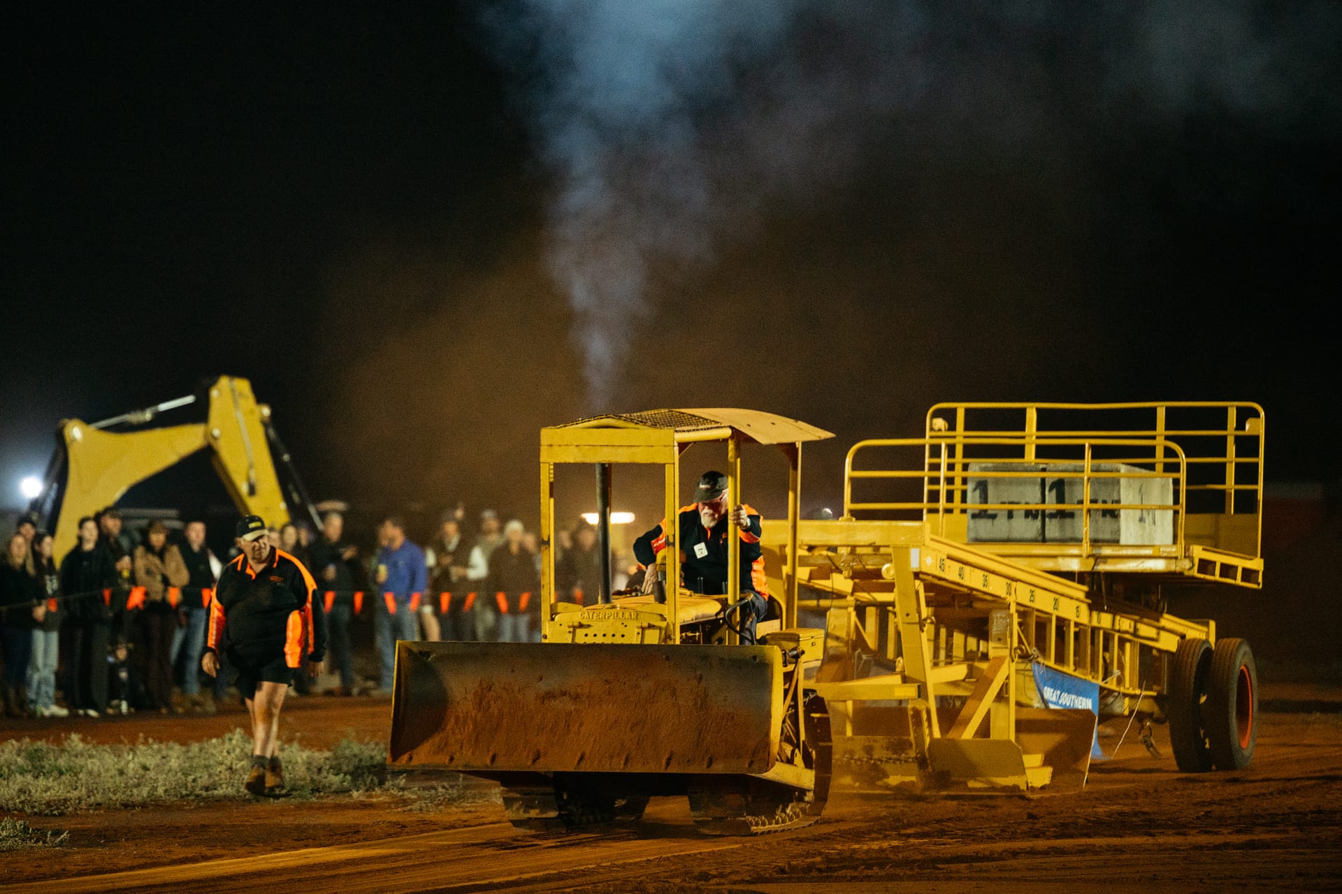 A yellow vintage Caterpillar crawler tractor pulls a heavy, weighted sled during a tractor pull event at night. Thick grey exhaust smoke billows upward from the tractor's vertical pipe. A driver wearing a dark jacket and cap steers from the open canopy cab. The tractor's front blade is lowered close to the dirt track. In the background, a crowd of spectators stands behind a safety line watching the event, and a large yellow excavator is parked in the distance under stadium lighting.