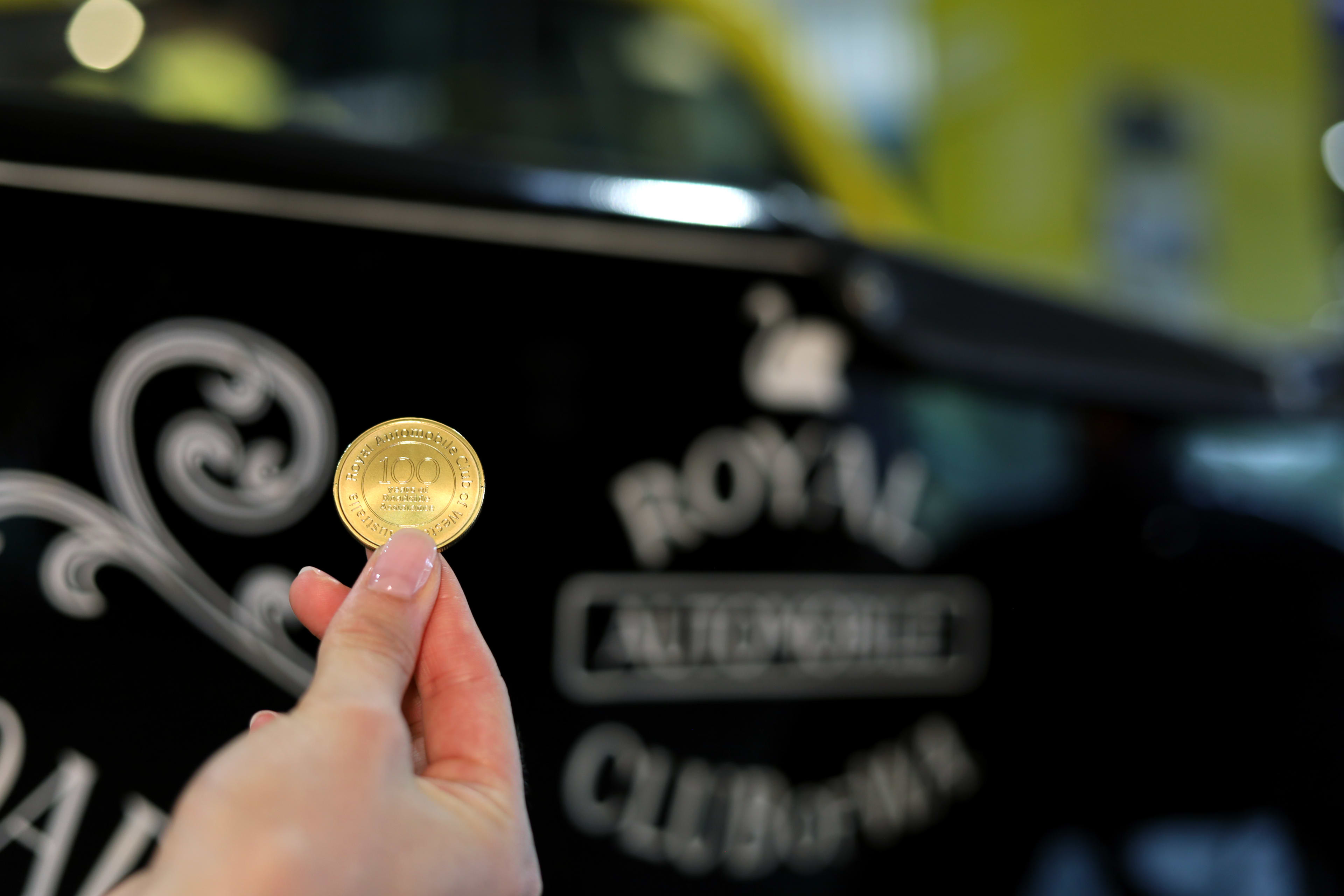 A person's hand holds up a gold-colored commemorative coin inscribed with "100" in front of a blurry, black vintage car featuring the white text "ROYAL AUTOMOBILE CLUB