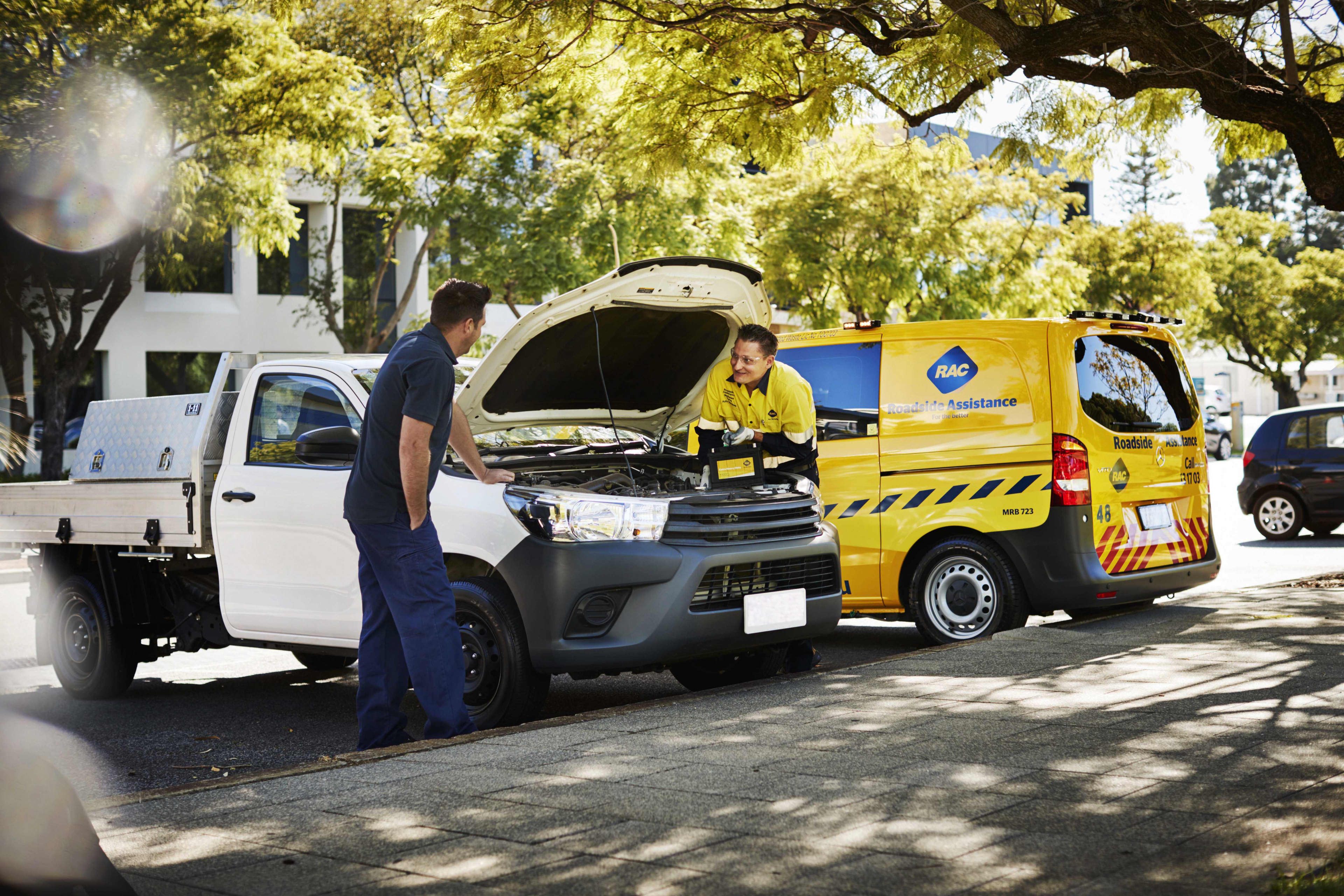 An RAC roadside assistance patrol technician in a high-visibility yellow uniform works under the open hood of a white utility truck on a tree-lined street, while the truck driver in blue workwear watches. A yellow RAC Roadside Assistance van is parked next to the truck.