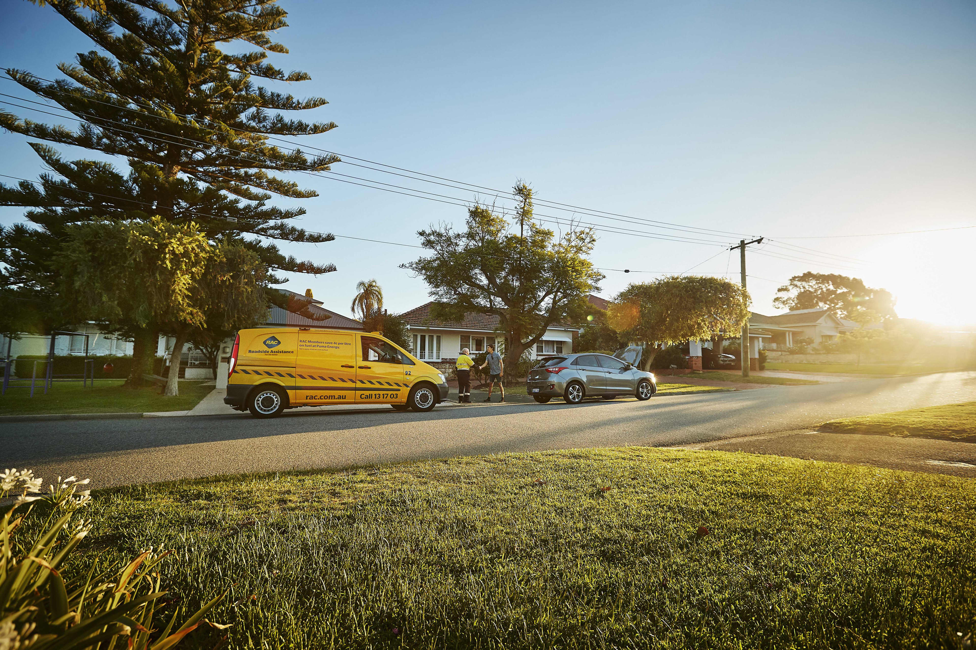 A wide shot of a suburban street under a bright sky, where a yellow RAC roadside assistance van and a gray car with its hood open are parked along the curb. In the background, an RAC technician shakes hands with a driver on the sidewalk next to suburban houses and green lawns.